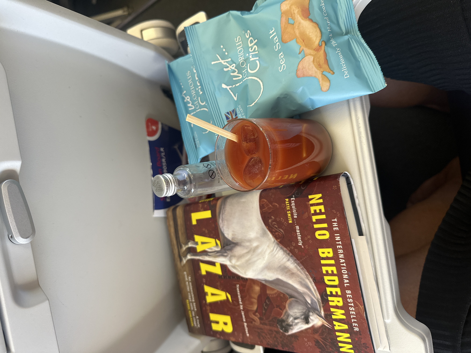 The image shows a tray table on an airplane, with a book, a drink, a small bottle, and two bags of crisps. The book is titled "LAZAR" and features an image of a horse. The drink appears to be a Bloody Mary, with ice cubes and a wooden stirrer. The small bottle is clear and has a silver cap, possibly containing water or another beverage. The bags of crisps are blue and labeled "Just... Glorious Crisps Sea Salt." The tray table is white and appears to be part of an airplane seat. The background is out of focus, but suggests the interior of an airplane cabin.