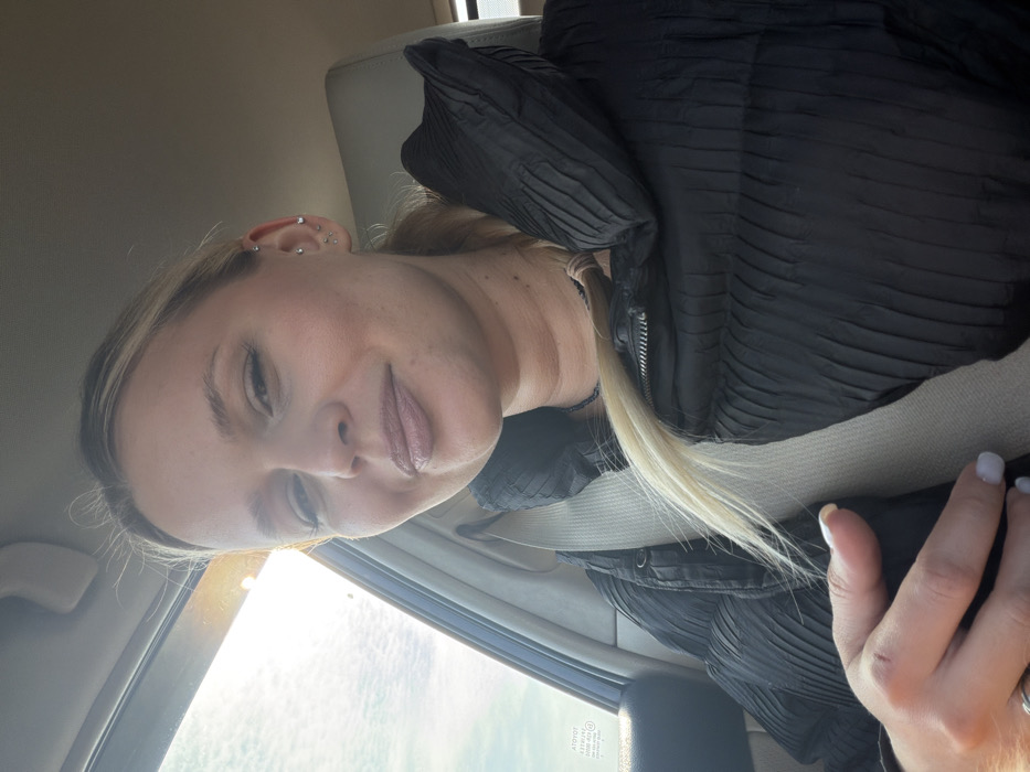 A young adult female is depicted in a selfie taken inside a vehicle in Povarenki, Russia. She has light skin, blonde hair styled back, and multiple ear piercings on her left ear. She is wearing a dark, possibly black, horizontally ribbed or quilted puffer jacket and has a light-colored seatbelt diagonally across her chest. Her left hand, with light-colored nail polish, rests near the lower part of the seatbelt. The woman is looking directly at the camera with a slight smile. Through the window on the left side of the vehicle, bright sunlight is visible against a partly cloudy sky, indicating daytime. Faint text, possibly "TOYOTA," is reflected on the window. The interior of the vehicle appears to be light-colored.