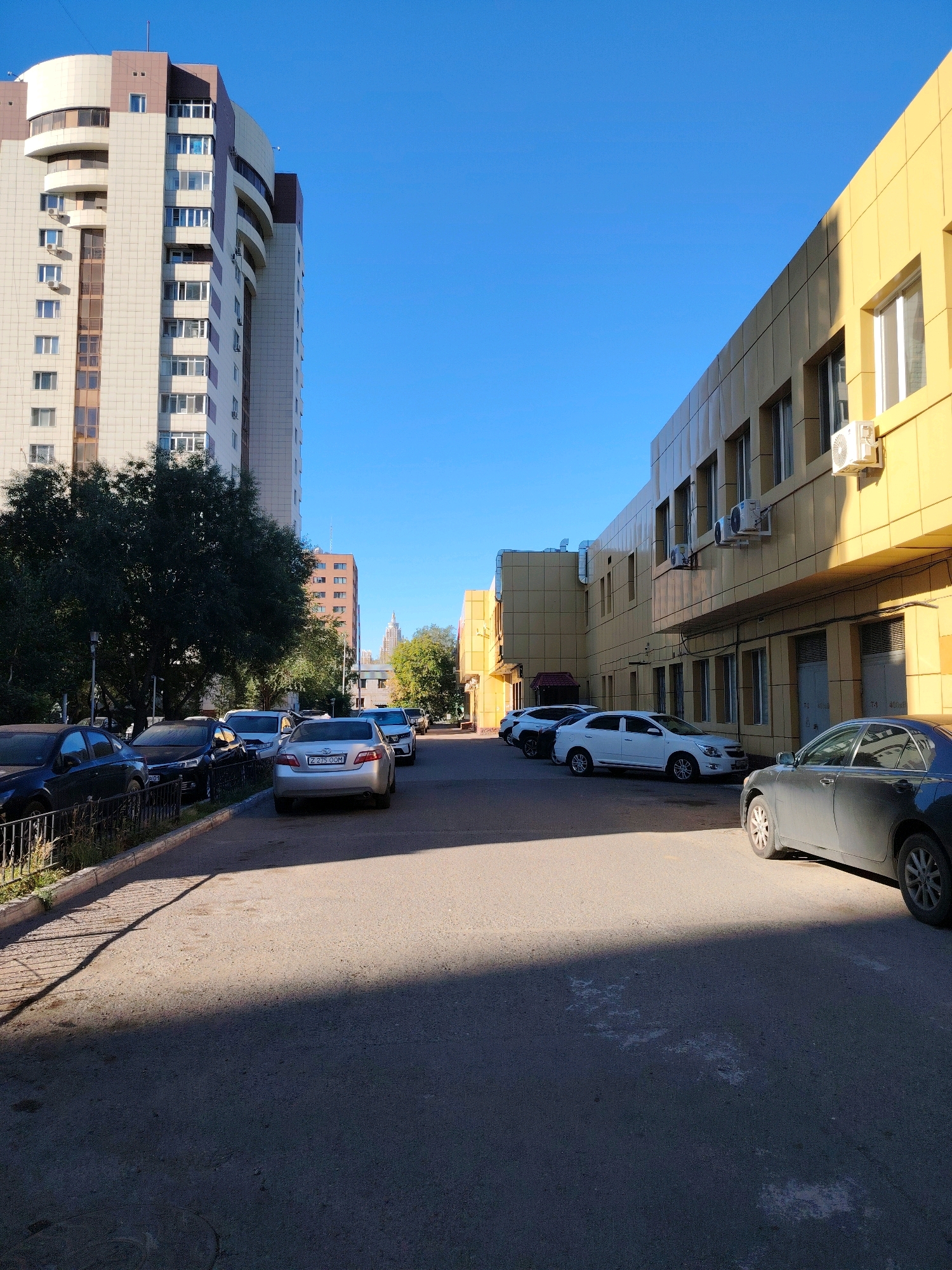 In a sunlit urban environment of Astana, Kazakhstan, a street scene unfolds with a focus on residential and commercial buildings and parked vehicles. Dominating the left side of the frame is a tall, multi-story residential building with a light-colored facade featuring numerous windows and balconies. Adjacent to it, a lower, yellow-clad commercial building stretches across the right side of the image, also with visible windows and outdoor air conditioning units.
Several cars are parked along the street and in a parking area. On the left, a black car is parked near a fence. In the foreground, a silver sedan with license plate "Z 275 OOM" is prominently visible. Further down the street, a white sedan and other vehicles are parked. Trees are interspersed throughout the scene, with a dense cluster of dark green foliage partially obscuring the base of the tall building. The clear blue sky suggests fair weather conditions. The ground is paved with asphalt, and a distinct shadow line cuts across the road, indicating the time of day and the position of the sun. The overall impression is of a typical day in a well-developed urban area.