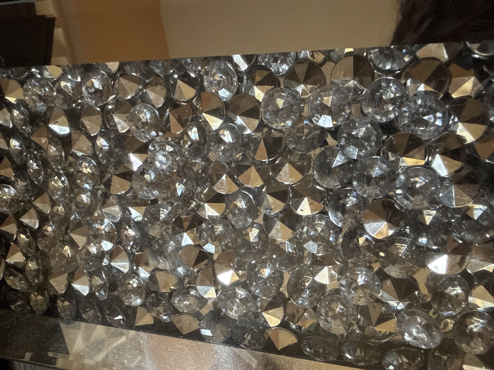 A close-up shot shows a surface densely covered with round, faceted crystals or rhinestones, appearing to be part of a decorative object. The facets of the crystals reflect light, creating a sparkling effect. The crystals are arranged haphazardly, varying slightly in size and orientation. At the bottom edge of the frame is a smooth, reflective surface, possibly a mirror or polished metal. The surrounding environment appears to be indoors, but further details about the location in Unknown, Unknown are not discernible from the image.