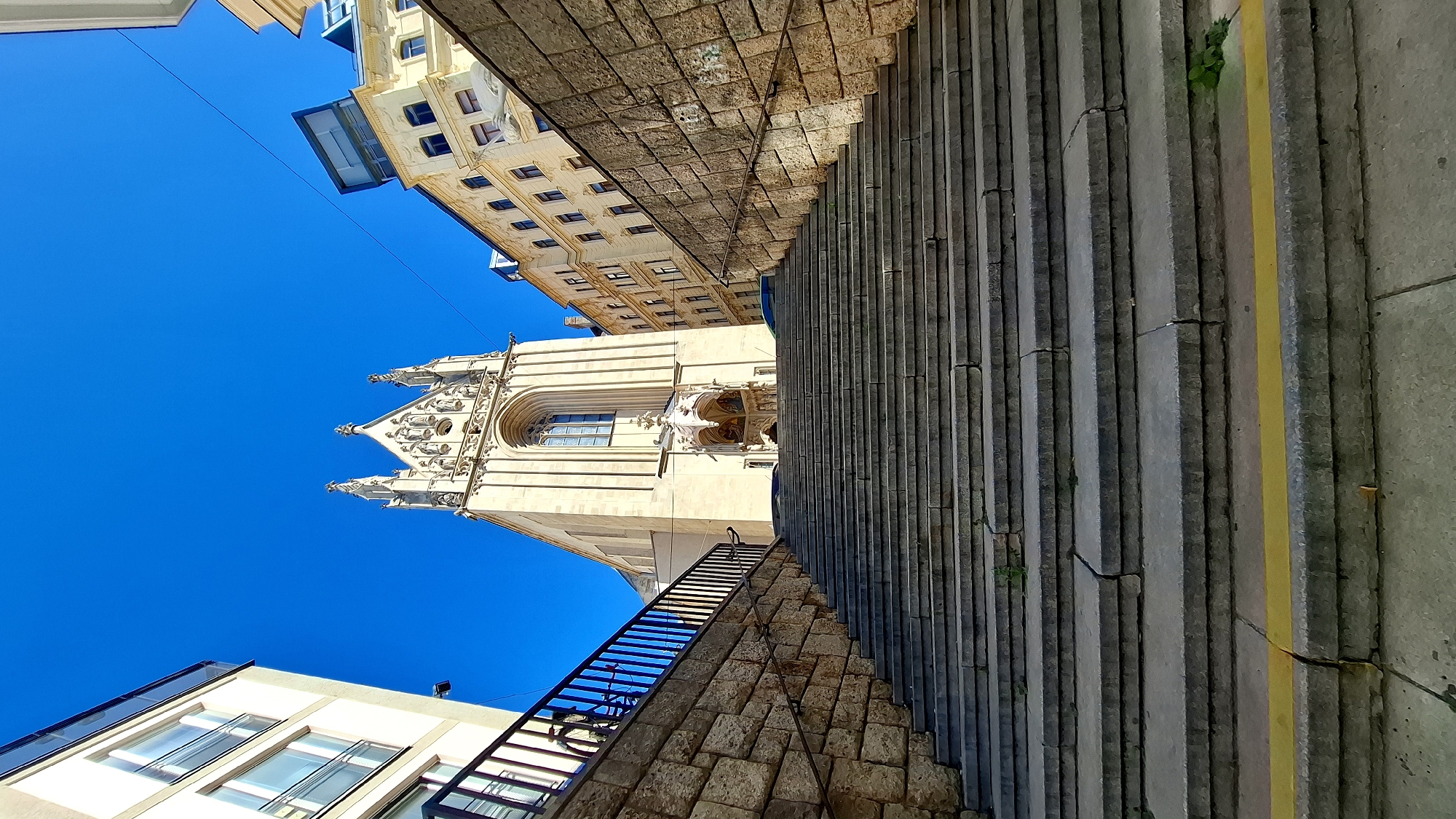 A low-angle shot captures a grand stone staircase leading up to the entrance of a Gothic-style church with a beige facade and a tall, ornate spire. The church is flanked by two buildings: a modern building with large windows and balconies on the left, and an older, light-yellow building with multiple windows and architectural details on the right. The sky above is a clear, vibrant blue, indicating a sunny day. The stone walls flanking the staircase are constructed from rough-hewn blocks. The steps themselves are wide and made of grey stone, some showing signs of wear and minor cracks. A yellow line is painted along the edge of one of the lower steps. The scene appears to be located in a European city, with architectural elements suggesting it could be Vienna, Austria. There are no people clearly visible in the immediate foreground, but a figure can be seen in the dimly lit entrance of the church. The overall impression is one of historical grandeur and an urban setting bathed in bright sunlight.