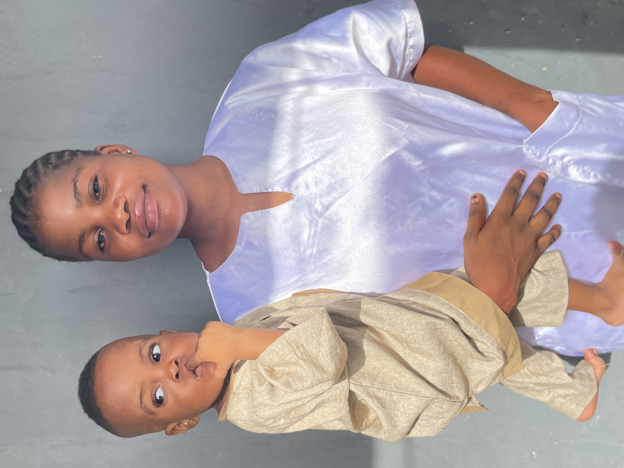 The image captures a Black woman and a young child, likely a mother and her baby or toddler, posed together on a flat, dark gray surface under bright, natural light in Badagry, Nigeria.
The woman is positioned closer to the top of the frame, dressed in a striking, reflective white long-sleeved tunic that shimmers in the sunlight. She has dark braided hair, a subtle nose piercing, and an earring. She looks directly at the camera with a gentle, faint smile. Her right arm is seen resting near the child, suggesting a protective or affectionate embrace.
Below her, the child is dressed in a textured, light beige long-sleeved garment, possibly a traditional outfit. The child has short, dark hair and wide, curious eyes, looking upwards towards the camera with one hand near their mouth. The plain gray surface provides a neutral background, highlighting the subjects. The bright illumination and distinct shadows indicate a sunny outdoor setting during daytime. The image conveys a serene and intimate moment between the two individuals.