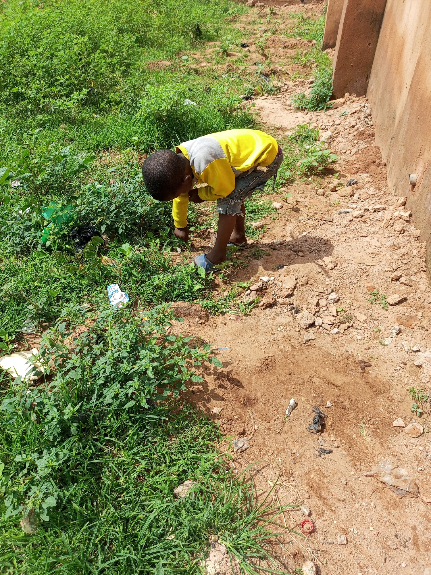 A young Black child, wearing a yellow and gray long-sleeved shirt and denim shorts, is bent over in a patch of dry dirt and scattered rocks next to a building with a textured, tan wall in Tempure, Nigeria. The child appears to be examining something on the ground. Lush green grass and weeds surround the dirt patch, with some scattered pieces of trash visible, including a white and blue wrapper, a plastic bottle cap, and what appear to be plastic bags. The ground is uneven and dusty.