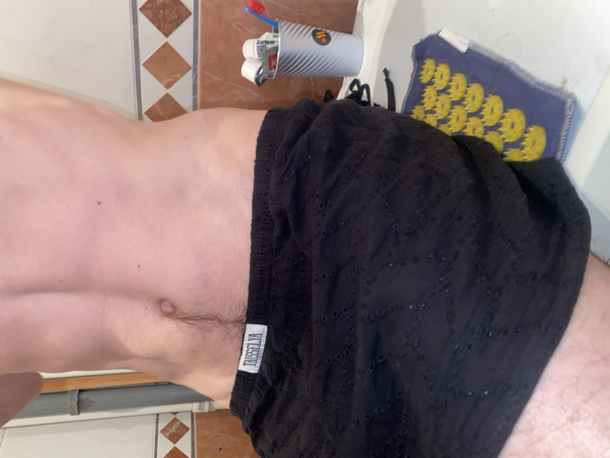 The image shows a person in a bathroom, wearing black shorts. A cup with a toothbrush and toothpaste sits near a sink with a blue and yellow mat. The bathroom has tile walls. There is no evidence to indicate that the location is Moscow, Russia.