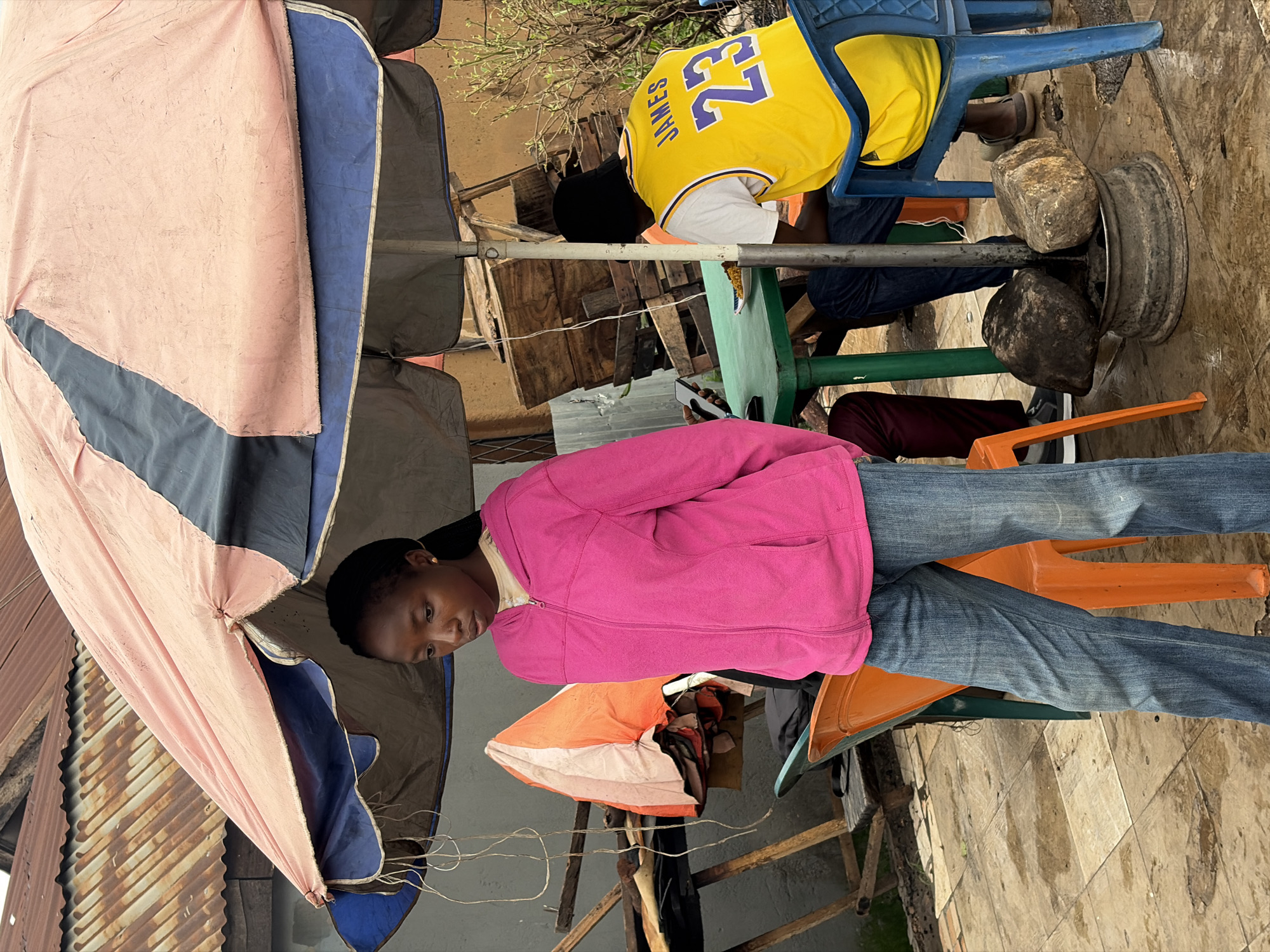 This image depicts an outdoor scene in Jos, Nigeria, likely a street-side eatery or gathering spot. A young woman in a bright pink hooded jacket and blue jeans stands in the foreground, looking downwards. She has dark braided hair and a small gold earring. Behind her, a large umbrella with pink and blue sections provides shade.
Under the umbrella, a green plastic table is set up with at least one plate of food. A person wearing a yellow basketball jersey with "JAMES" and the number "23" on the back is seated at the table. Another person, mostly obscured, is also at the table, holding a mobile phone. Several plastic chairs in orange and blue are scattered around the table.
The environment appears somewhat informal, with a corrugated metal roof visible in the background. Wooden crates and other assorted objects are stacked behind the table. The ground is paved with tiles and appears wet, suggesting recent rain. The overall impression is a casual, everyday scene in a Nigerian urban setting.