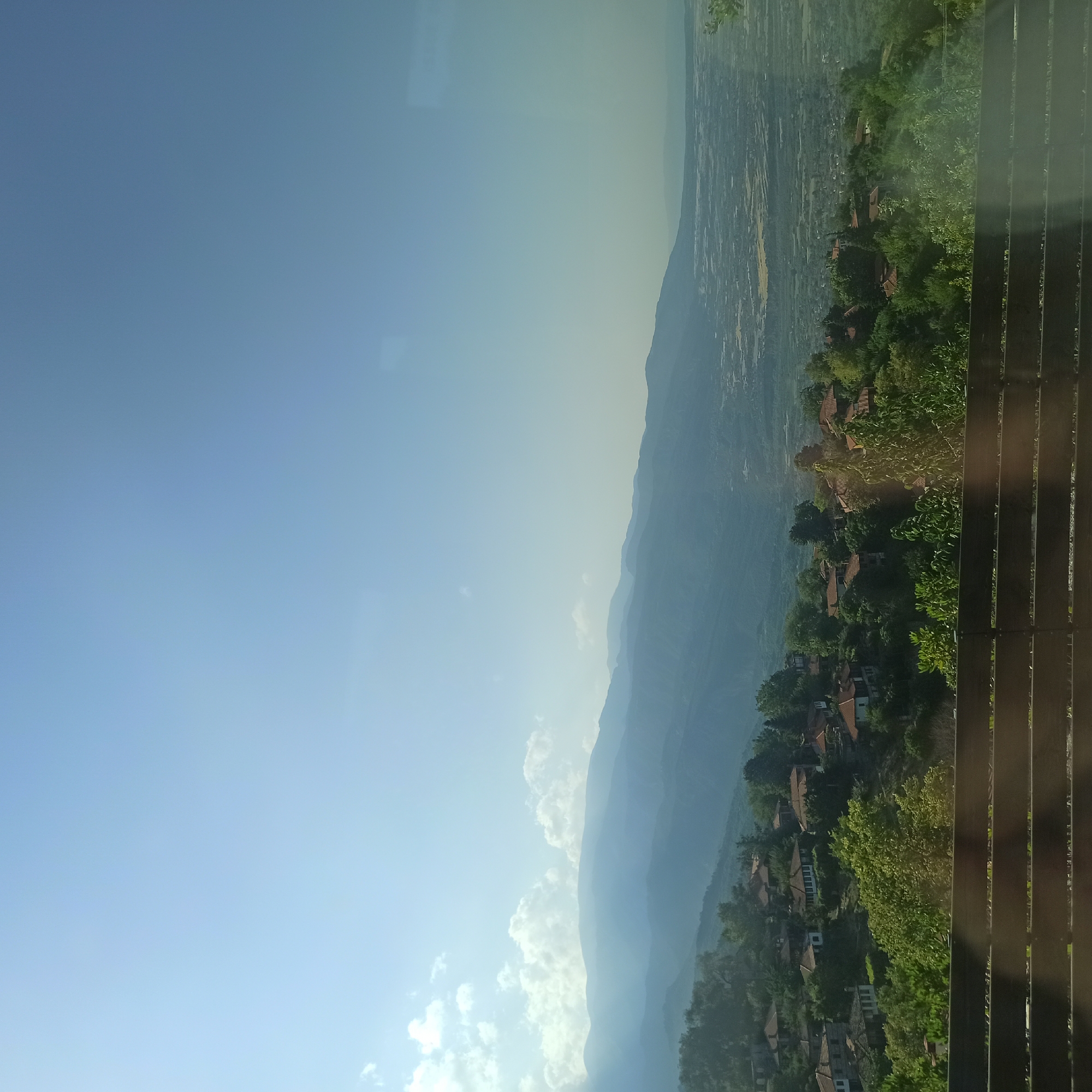 This is a vertical, eye-level, medium-shot photograph taken from a high vantage point. The scene is a sprawling landscape with layers of mountains receding into the distance under a clear blue sky. The sky is bright and appears to be from the daytime, possibly morning or afternoon, with minimal cloud cover, save for a few wispy clouds visible near the horizon. The light suggests a sunny day.
In the foreground, to the right, there is a wooden railing, which appears to be part of a balcony or viewing platform. The railing consists of dark brown, vertical slats. Beyond the railing, the landscape unfolds. A village or settlement is nestled into the hillside, characterized by numerous houses with terracotta tile roofs, interspersed with dense green trees. The houses are scattered and follow the contours of the terrain, creating a picturesque view. The vegetation is lush and varied, with a mix of trees and shrubs.
The background is dominated by undulating mountain ranges that fade into a hazy blue due to atmospheric perspective. The distant valley floor is visible between the mountain ranges, appearing as a patchwork of fields or possibly another settlement.
The overall mood is serene and expansive, offering a breathtaking view of a rural or semi-rural mountainous region. There is no visible text in the image. The image appears to be a landscape shot, potentially taken from a tourist viewpoint or a residential property with a scenic overlook, possibly in Neos Panteleimonas, Greece, given the context.