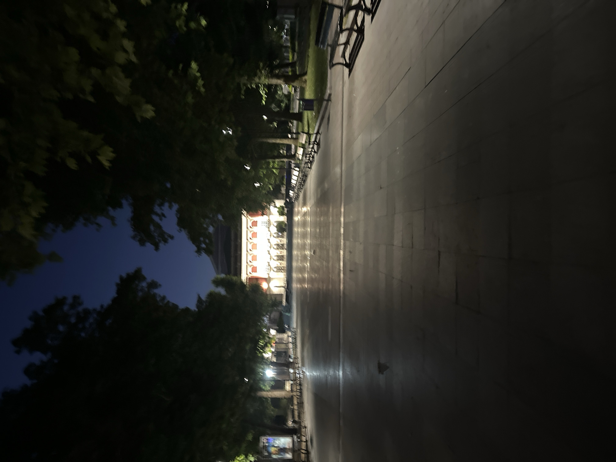 The image depicts an outdoor public space at night, likely a plaza or park area, in Ruse, Bulgaria. The scene is illuminated by artificial lighting, casting reflections on the wet pavement. Rows of park benches line the pathway, some appearing unoccupied. Tall trees with dark foliage border the walkway, their branches forming a canopy overhead against the dark blue night sky. In the distance, a brightly lit building with ornate architecture stands prominently, its windows and facade illuminated, suggesting it might be a cultural institution or government building. The overall atmosphere is calm and empty, characteristic of a quiet urban environment after dark.