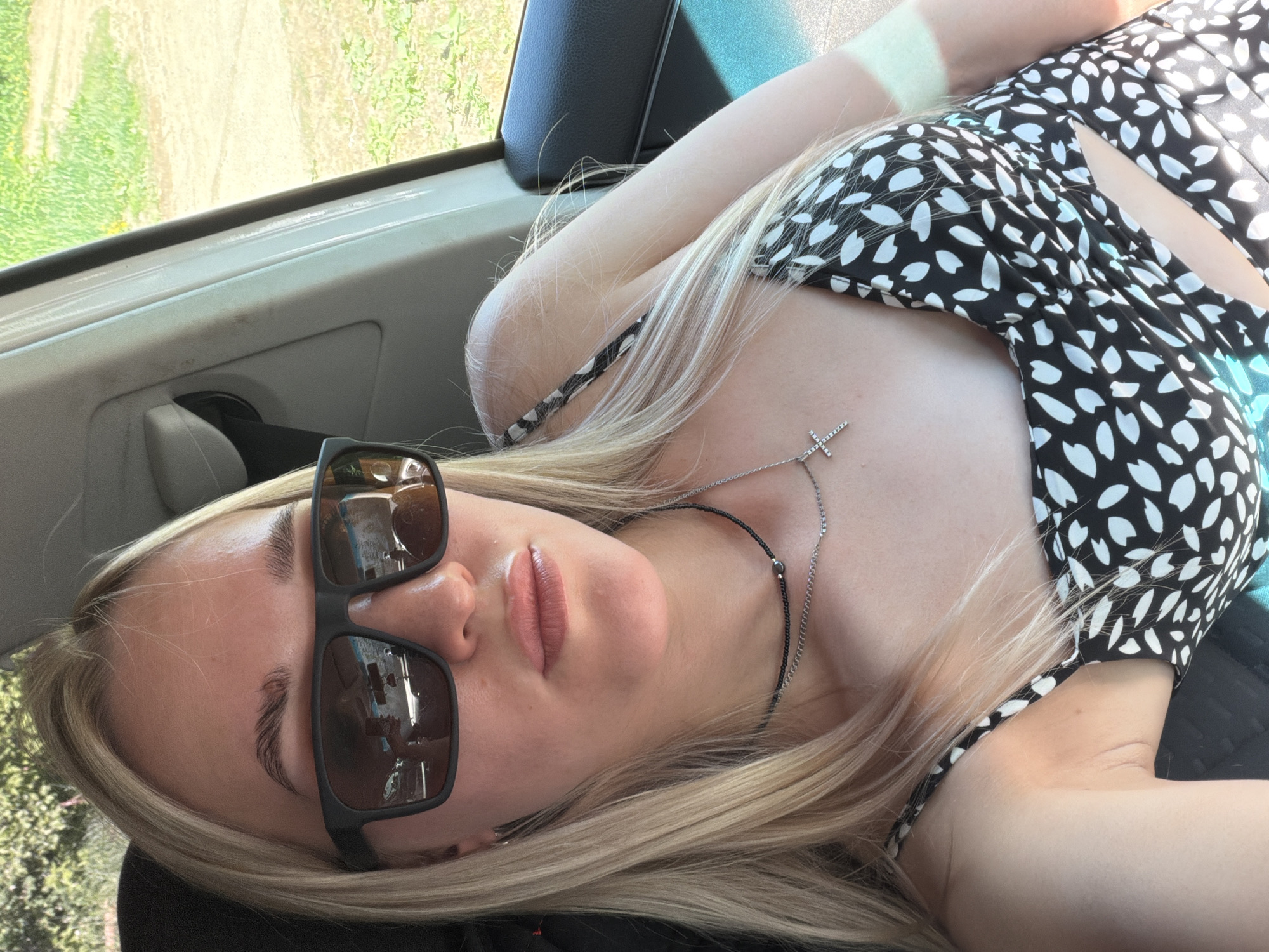 The image shows a blonde woman wearing sunglasses and a black and white patterned top. She is lying down in what appears to be a car, with the window and door visible in the background. The reflection in her sunglasses suggests an urban environment outside the vehicle. She is wearing two necklaces, one with a cross pendant and the other a beaded necklace. The location in the city is Unknown, Unknown.