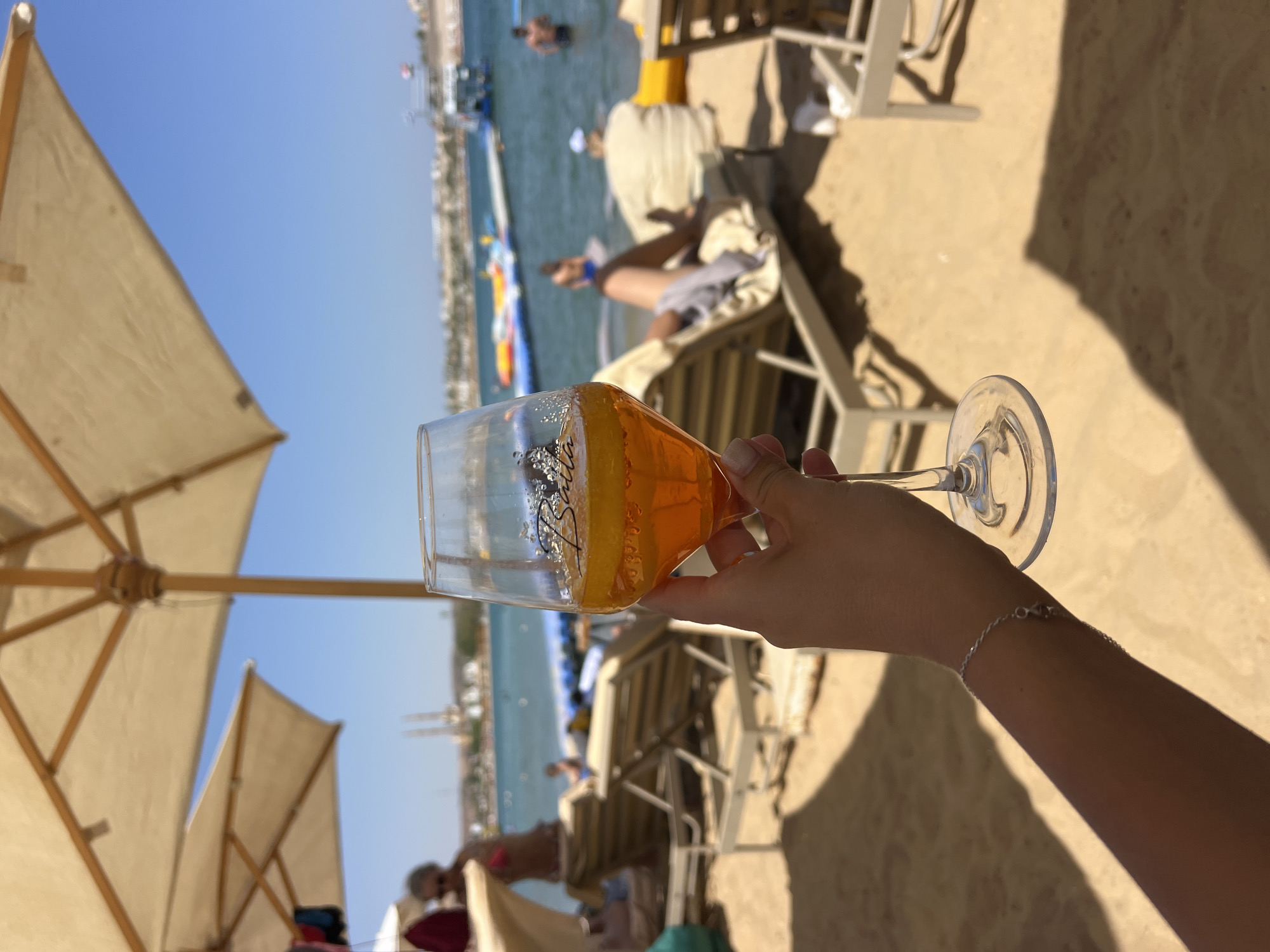 A hand holds a wine glass filled with an orange beverage, likely Aperol Spritz, on a sunny beach. In the background, beach umbrellas shade empty lounge chairs. Beyond the chairs, people are visible in the turquoise water, some swimming and others on floating water toys. A distant shoreline with buildings and structures can be seen under a clear blue sky. The scene is characteristic of a resort beach in Sharm El Sheikh, Egypt.