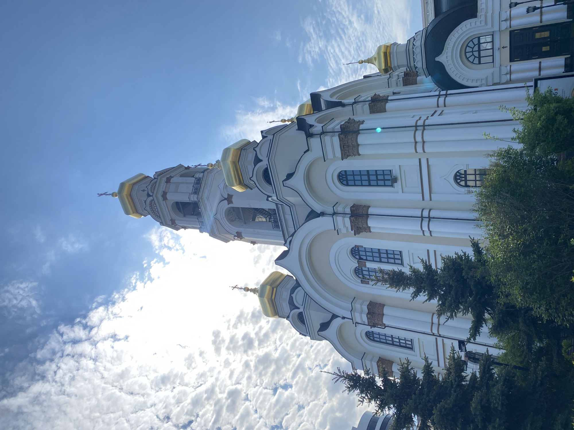 The image shows the Church on Blood in Yekaterinburg, Russia, viewed from a low angle. The church, primarily white with golden domes, reaches towards a blue sky with scattered clouds. Sections of the facade are visible, including windows and decorative elements. Green trees are visible in the foreground.