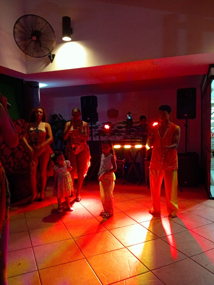 The image depicts an indoor entertainment venue in Playa El Agua, Venezuela, under stage lighting characterized by dominant red and orange hues on the tiled floor and subjects. Six individuals are present: three adult women, two children, and one adult male. On the left, two adult women wearing sequined two-piece performance costumes are visible, with one having her hands clasped. A third adult female is partially obscured on the far left. In the foreground, two children are observing the performers: a young girl in a floral dress stands on the left, and an older girl in a light-colored top and pants stands on the right. On the far right, an adult male, dressed in a bright orange/yellow two-piece costume, stands facing towards the center. In the background, a DJ booth with mixing equipment is visible, illuminated from below, flanked by two black speaker cabinets. A male individual is positioned behind the DJ booth. Overhead, a black wall-mounted oscillating fan and a black cylindrical wall-mounted spotlight are affixed to the light-colored ceiling. The scene suggests a live performance or show.