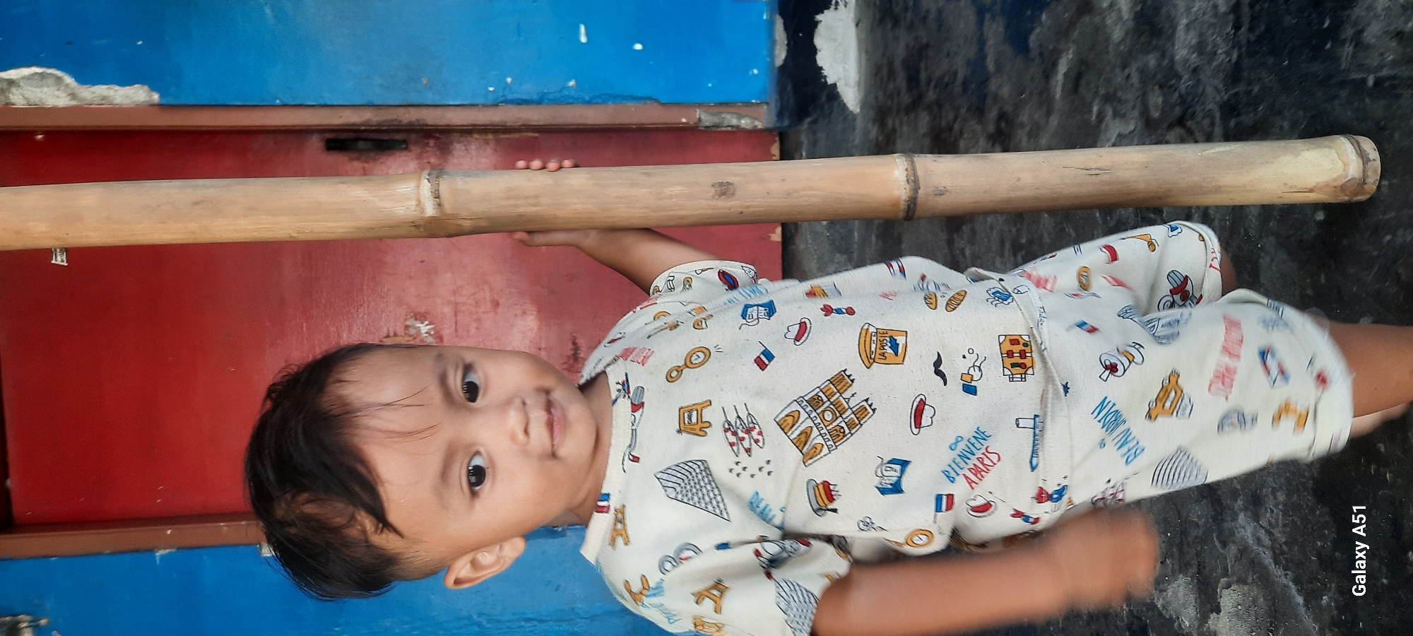 A young child, likely a toddler, stands in front of a red and blue painted wall, holding onto a large bamboo pole. The child is wearing a light-colored t-shirt and shorts with a pattern of various small images, including buildings, flags, and text. The child's hair is dark and styled with a visible undercut. The background suggests an outdoor or semi-outdoor setting, possibly a doorway or entrance, with a rough, textured floor. The lighting is natural and appears to be daylight. The image is tagged "Galaxy A51," indicating it was captured with a Samsung mobile device. The location is identified as Selopuro, Indonesia.