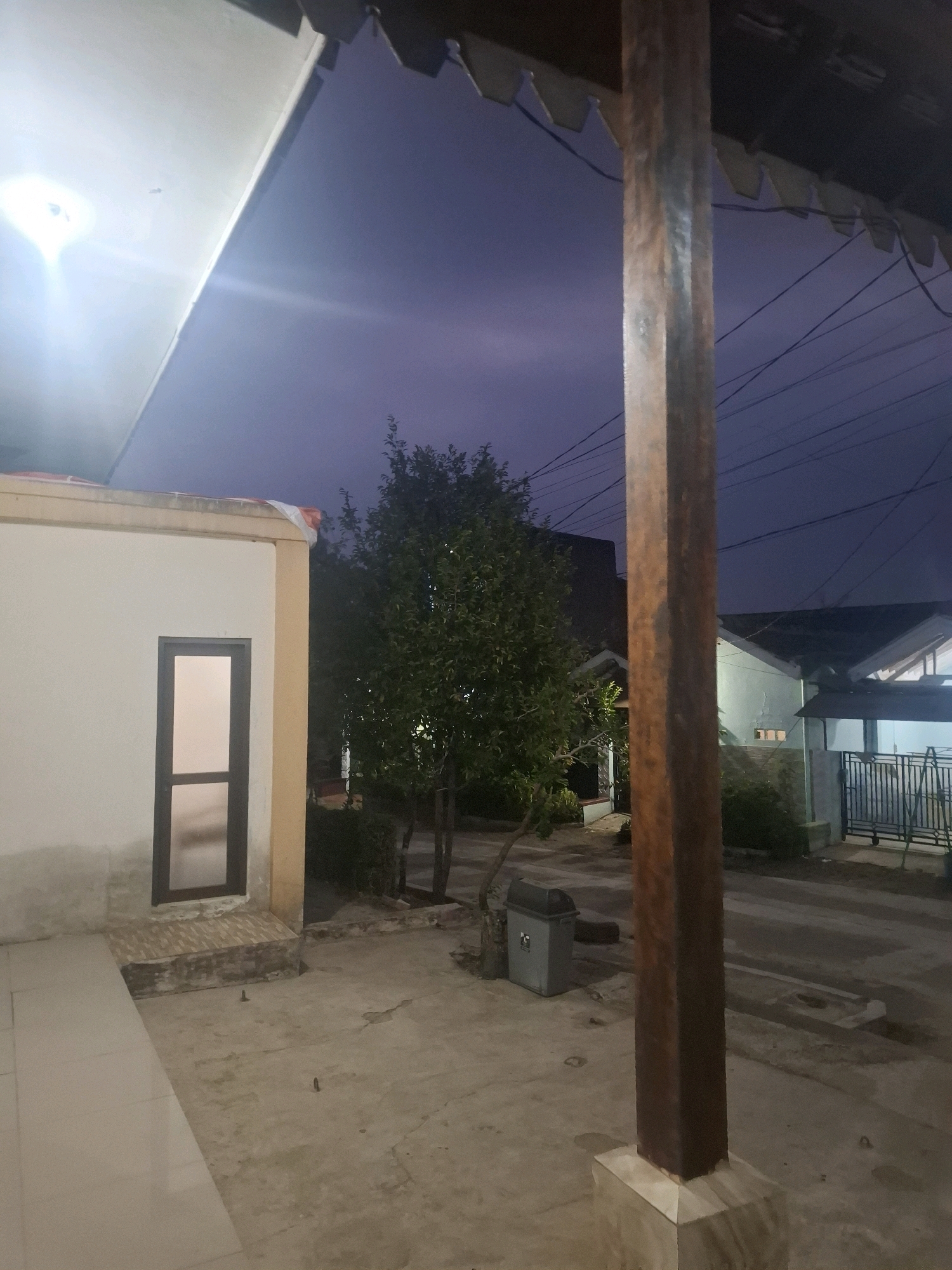 The image depicts an outdoor scene at dusk or early night in what appears to be a residential area in Ciheulang Tonggoh, Indonesia. A building with a beige facade and a dark-framed window is visible on the left. A large, dark wooden post stands prominently in the foreground on the right, supporting a portion of a dark-colored roof structure with visible scalloped edges and electrical wires running overhead. Behind the building and post, trees with dark green foliage are present, and further back, houses with lighter colored walls and roofs can be seen. A gray trash bin is situated near the base of a tree. The ground is paved with concrete. The sky is a deep, dusky blue, suggesting twilight. There are no people or animals visible in the image. The overall impression is a quiet, domestic exterior setting.