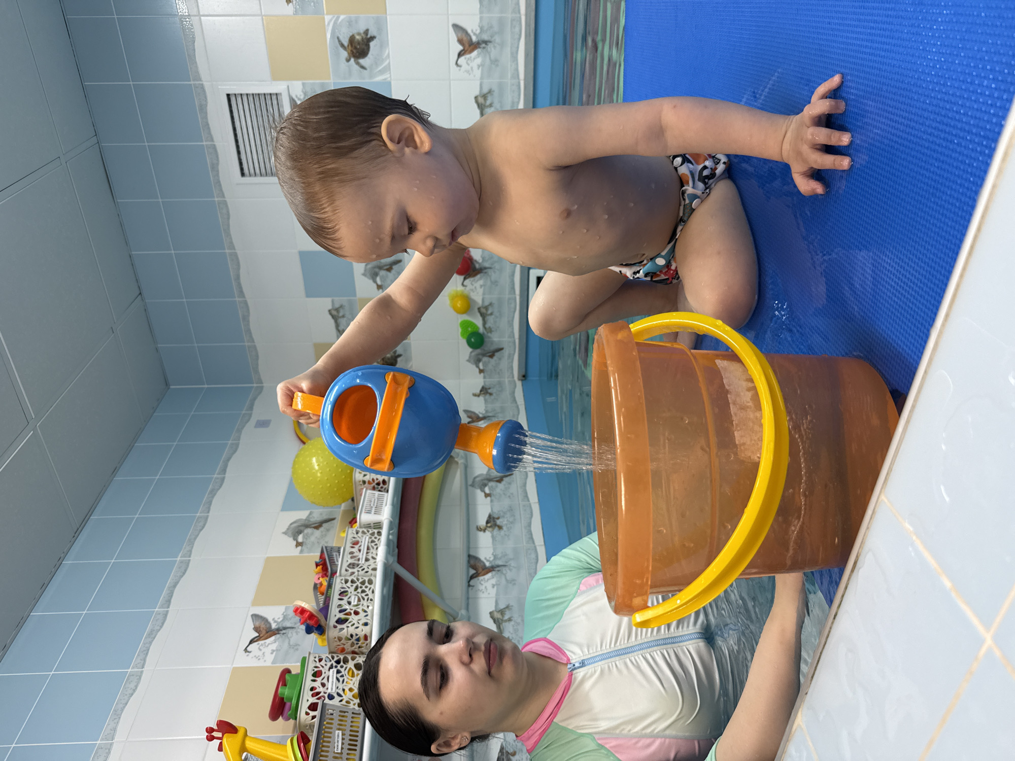 A young child is kneeling on a blue mat, pouring water from a blue and orange watering can into a clear orange bucket held by a woman. The woman is wearing a white and pastel rash guard. The scene appears to be indoors, possibly in a swimming pool area, as there are tiled walls with decorative bird patterns, pool markings, and toys in the background. The setting is Rusanovo, Russia.