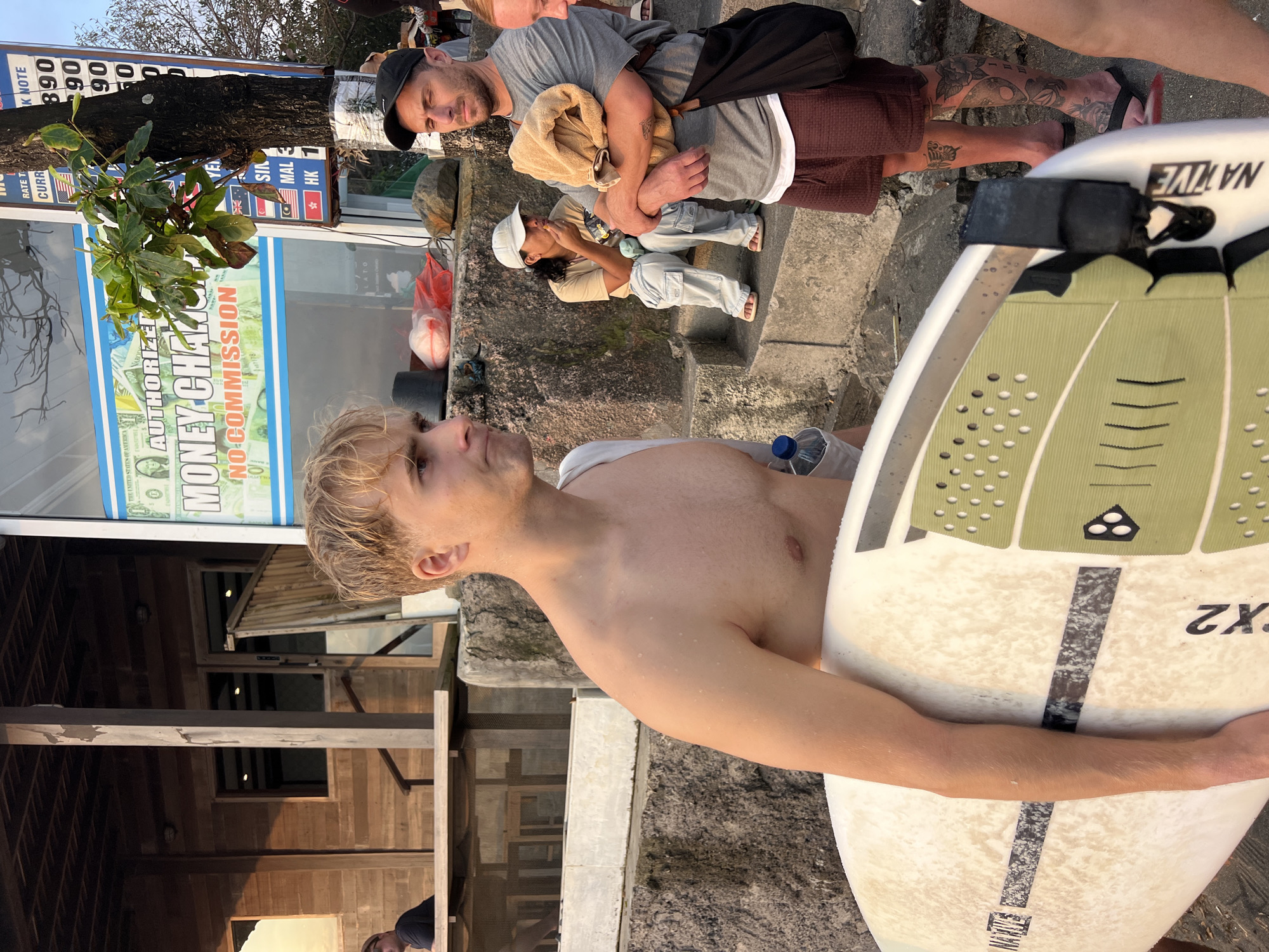 The image depicts a street scene in North Kuta, Indonesia. In the foreground, a shirtless young man with blond hair holds a surfboard. To his right, a man wearing a black baseball cap and a grey t-shirt holds a folded towel and has a black bag slung over his shoulder. Behind them, a woman in a light-colored cap and light-colored pants is seated and eating. In the background, a wooden building with a porch and windows is visible, along with a money exchange booth displaying currency rates. The environment appears to be outdoors, with concrete stairs and a tree with signage.