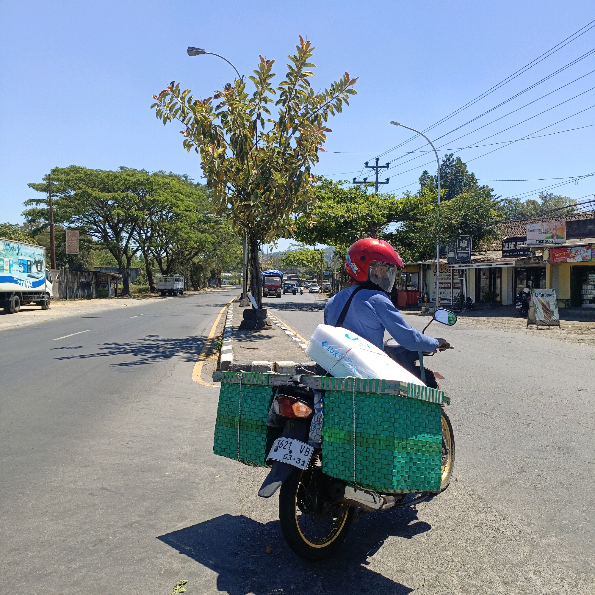 A person wearing a blue long-sleeved shirt and a red helmet is riding a motorcycle on a paved road. The motorcycle has a large green woven basket attached to the rear, which contains white items. The rider is looking to the right. The road is lined with trees and buildings. In the background, there are other vehicles on the road, including trucks and cars, and utility poles with wires. The sky is clear and blue. The scene appears to be in a commercial or residential area with a mix of natural and man-made elements. The license plate on the motorcycle reads "3621 VB G3 31". There is a sign on a building in the background that says "MIE AYAM HAYSO".