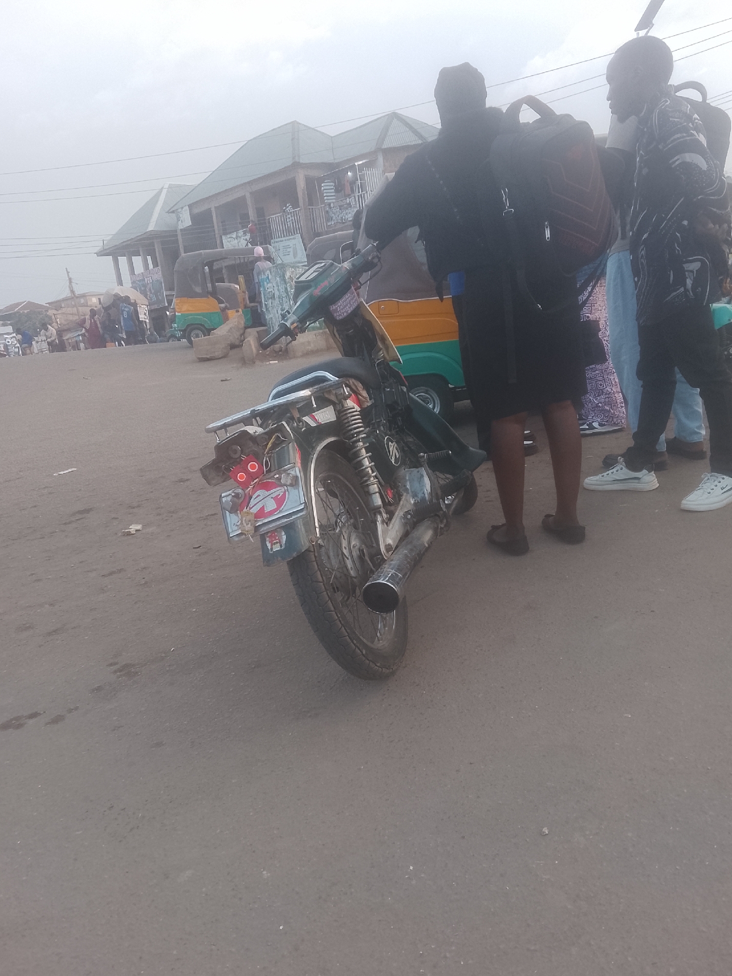 The image depicts a street scene in Gwong, Nigeria. A motorcycle with a damaged rear and license plate is parked on the asphalt. Several people stand nearby, partially obscuring the view. One person is wearing a black outfit and carrying a backpack. Another person, wearing a patterned shirt and black pants, stands next to them. In the background, a building with multiple floors and a green roof is visible. Also in the background is a parked tricycle. The weather is overcast.