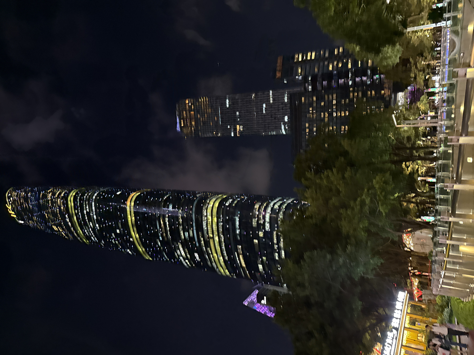 At night, the image captures a view of the city within the Tianhe District of Guangzhou, China. Prominently featured are illuminated skyscrapers against a dark sky, specifically, the Guangzhou Circle Mansion and Guangzhou CTF Finance Centre. Trees and other plant life intersperse the cityscape, adding a natural element to the urban scene. Below the skyscrapers, there are the illuminated storefronts and public spaces, indicating pedestrian activity.