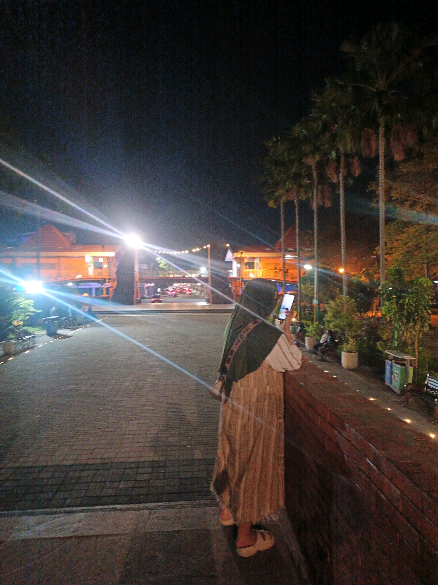 The image depicts a nighttime scene in front of the Trowulan Museum in Mojokerto, Indonesia. A person wearing a hijab and a long, striped dress is standing and looking at their phone. They are leaning against a brick wall. Palm trees are visible in the background, and the museum building, illuminated by lights, is to the left. Streetlights and car headlights are also present. The ground is paved with interlocking bricks. The overall ambiance suggests a warm evening.