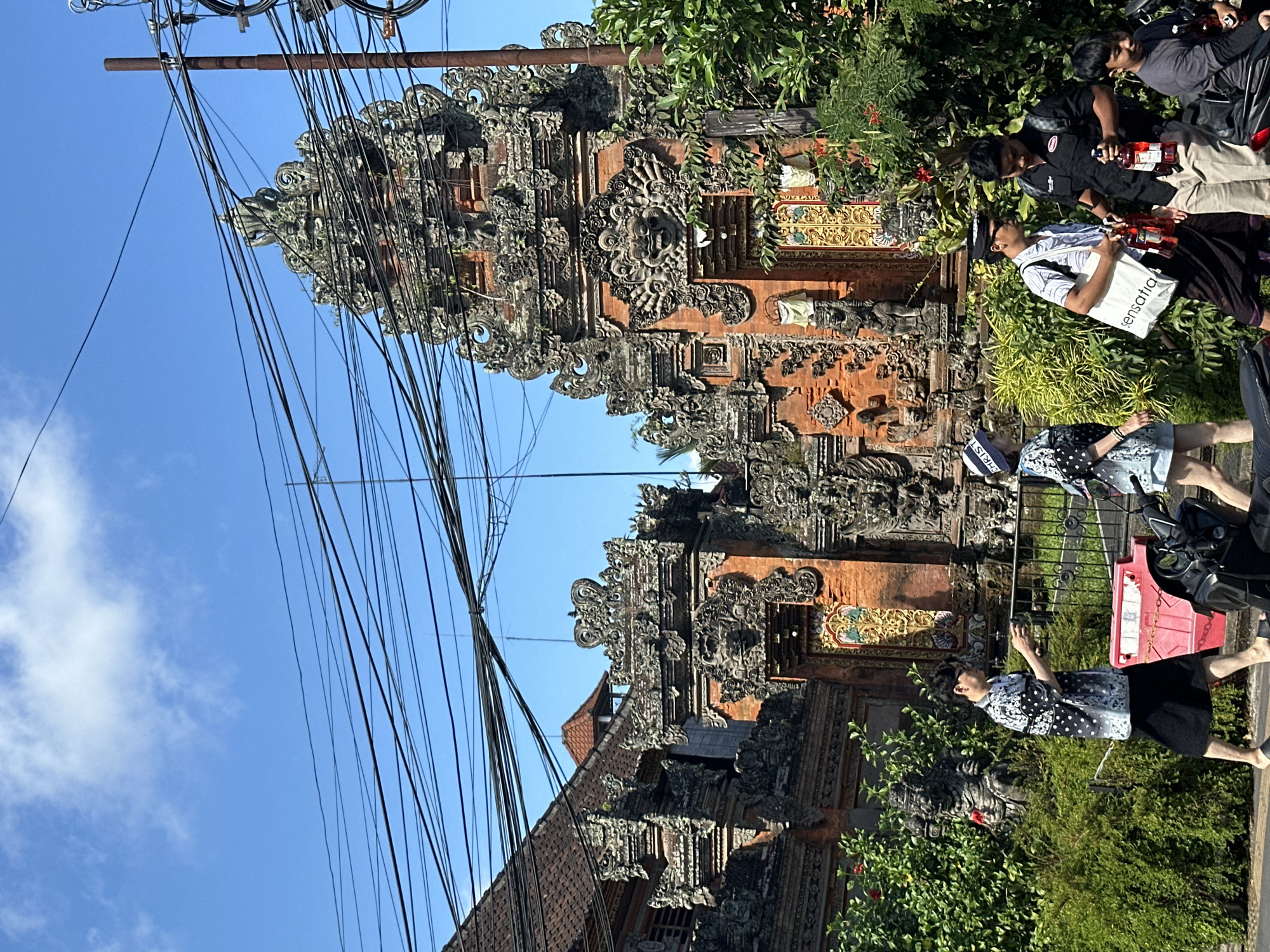 In the city of Ubud District, Indonesia, a street scene unfolds under a clear blue sky punctuated by a few white clouds. Prominently featured are intricate Balinese temple gates, adorned with ornate stonework and orange brick, partially obscured by a dense network of overhead electrical wires. To the left, a portion of a red-tiled roof and another section of a temple structure are visible.
In the foreground, a group of people are walking along the street. A man in a black and white patterned shirt and a black skirt-like garment is walking from left to right, holding a small item in his hands. To his right, a woman wearing a blue and white patterned outfit and a white hat is also walking from left to right. Further to the right, two men are engaged in conversation. One man, wearing a striped shirt, a dark sarong, and a cap, holds a white tote bag. The other man, dressed in a black shirt, is holding two bottles of red liquid. A young boy in a grey t-shirt is seated on a motorcycle to the far right, also holding a red bottle.
The area is bordered by lush green foliage and hedges. A red plastic barrier is placed on the ground near the center of the image, and several motorcycles are parked along the street. The overall impression is a vibrant street scene in a culturally rich environment, with a mix of traditional architecture and modern elements.