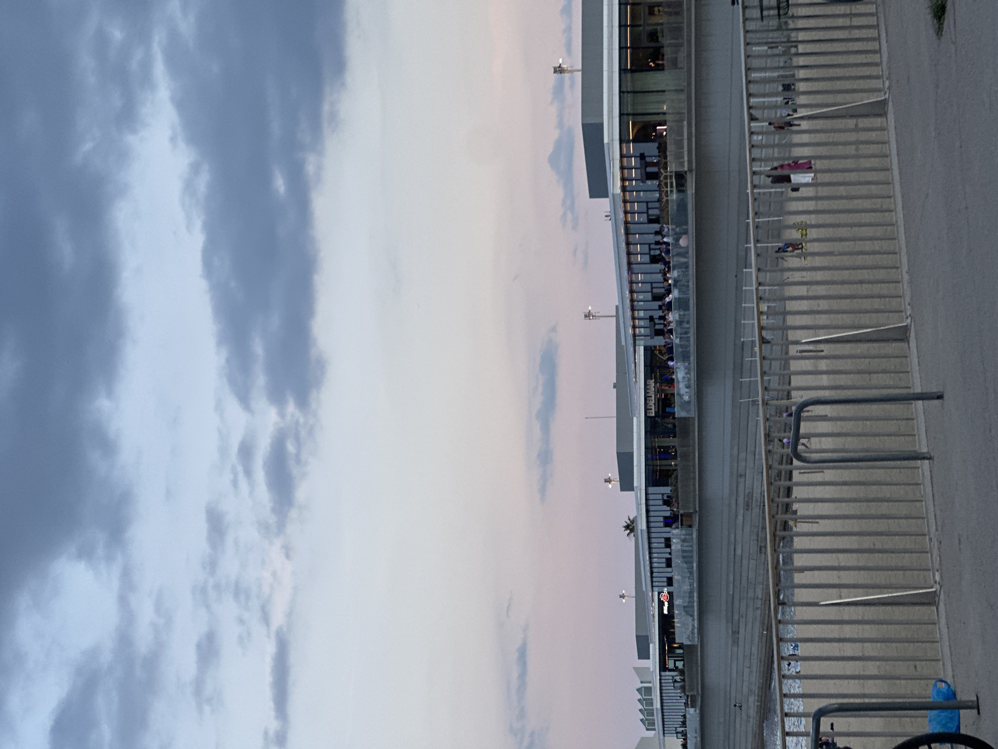 The image captures a waterfront scene in Barcelona, Spain, likely near the Parc del Fòrum area, characterized by modern architecture and a beach. In the foreground, a metal fence with vertical bars separates a paved walkway from a sandy beach. A blue plastic object and a bicycle wheel are visible on the walkway to the left. The beach itself has a few scattered figures.
Behind the beach, a large concrete structure with tiered seating descends towards the water. Above this, a sleek, modern building complex runs horizontally across the frame. This building features extensive glass facades, revealing people gathered inside. Signs are visible on the building, including one that reads "EDELMAR". Lights are illuminated within the building, suggesting it is evening or dusk. The sky above is a blend of twilight colors, with soft pinks and grays, and scattered dark clouds. The overall impression is of a contemporary urban space designed for leisure and entertainment, illuminated by artificial lights against the natural light of the fading day.
