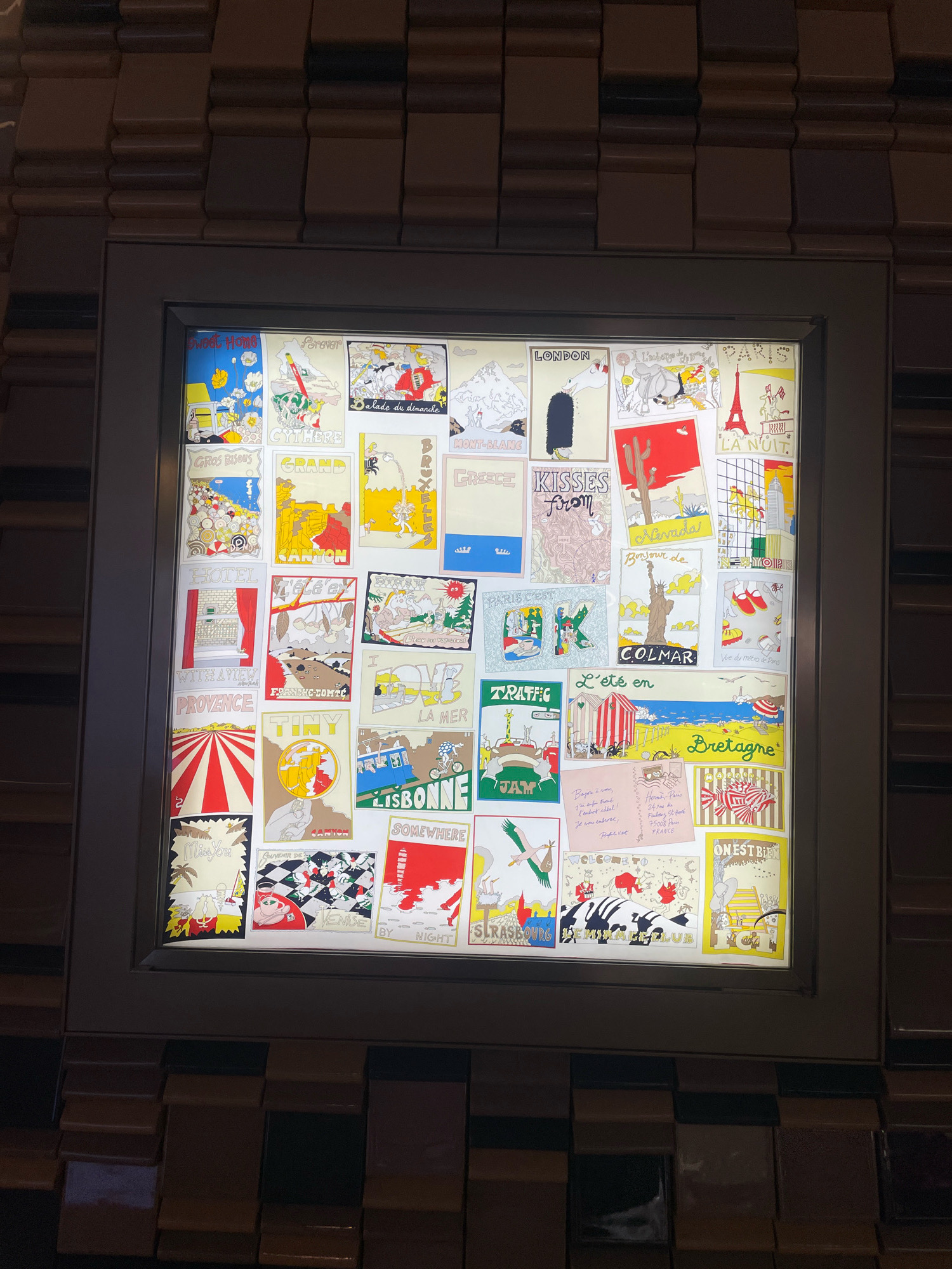 The image shows a framed display of small, colorful postcards or art prints, arranged in a grid-like pattern on a lighted panel. The artworks feature various whimsical illustrations, many depicting travel destinations, landmarks, or scenes with text in French and English. Examples include depictions of Paris with the Eiffel Tower, London, New York, and various landscapes. The display is mounted on a wall covered in textured, dark brown panels that resemble stacked, square tiles with varied depths. The lighting from the panel illuminates the artwork and casts a soft glow. The setting appears to be indoors, possibly a hotel lobby, restaurant, or retail space. Based on the visual style of the display and the surrounding decor, this scene is likely located in Doha, Qatar.