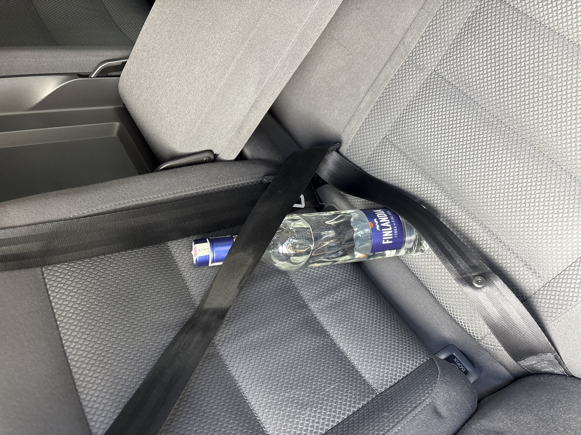 The image shows a bottle of Finlandia vodka secured by a seatbelt on the back seat of a car. There are no people visible. The car seat is gray with a textured surface. The bottle is approximately half full of a clear liquid. The environment is limited to the car's interior. There is no evidence of the location being in Dolno Drianovo, Bulgaria, or any other specific location.