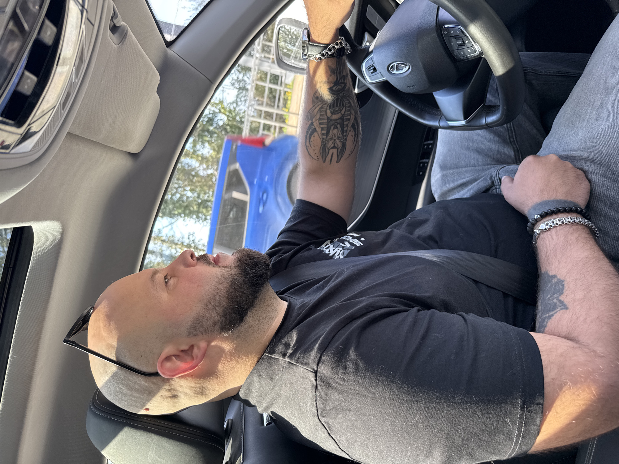 A bald, bearded man with a tattoo on his left arm is shown from inside the driver's seat of a vehicle. He's wearing a black t-shirt and grey jeans, and a seatbelt. A silver bracelet adorns his right wrist, and sunglasses are tucked behind his ear. He is holding the steering wheel of a Chery automobile. Outside the vehicle, a blurred blue and red object is visible. The location cannot be determined as Sochi, Russia based on visual information.