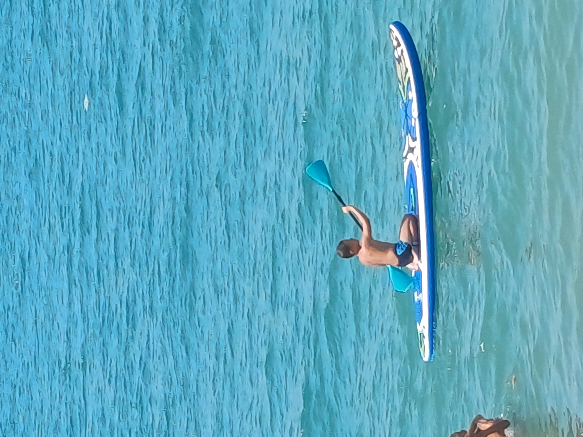 A young boy is kneeling on a paddleboard in the turquoise waters of the Black Sea, near Sochi, Russia. He is holding a blue paddle and appears to be paddling. The paddleboard is white with blue and colorful designs. The water is choppy, with small waves. In the foreground, to the left, another person is partially visible in the water. The sky is a pale blue.