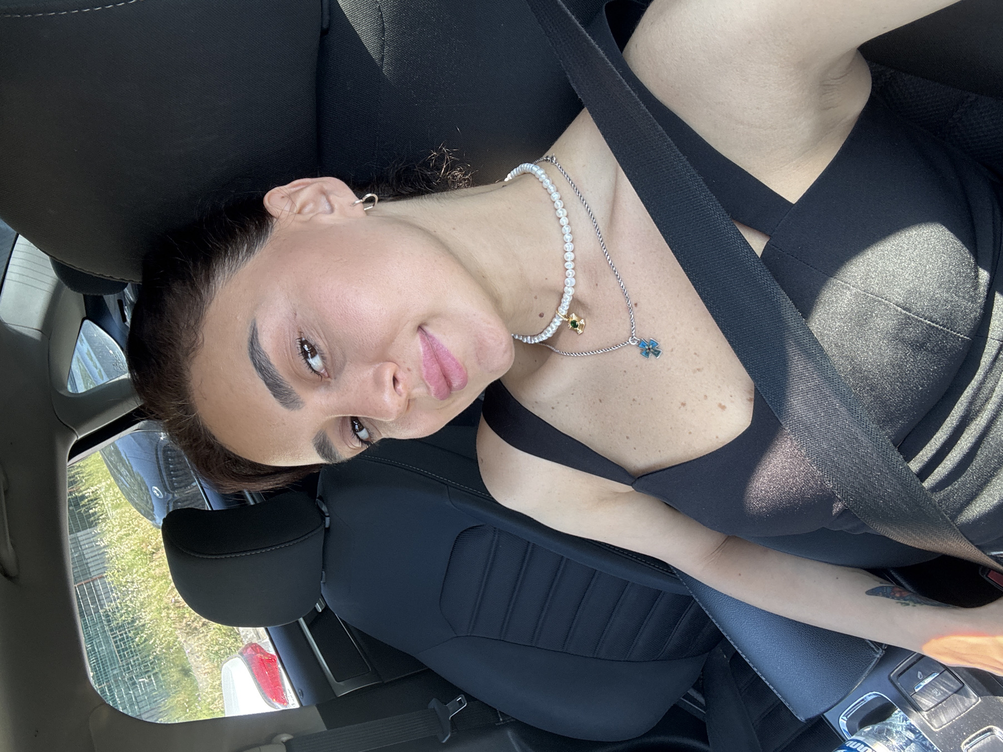 A medium shot captures a young woman with dark hair pulled back and a light tan complexion, seated in the passenger seat of a car. She is looking towards the camera with a pout and has light makeup on, including eyeliner and blush. Her outfit consists of a black tank top, a pearl necklace with a green charm, and a silver necklace with a blue cross pendant. She is wearing a small hoop earring in her left ear and has a black seatbelt fastened across her chest. A partial view of a car interior is visible, including the black seat, part of the dashboard with a bottle of water, and a car console. Through the car window, a black BMW sedan is parked in a grassy area with a chain-link fence in the background. The scene appears to be outdoors in daylight, possibly in a parking lot or along a roadside in Tbilisi, Georgia.