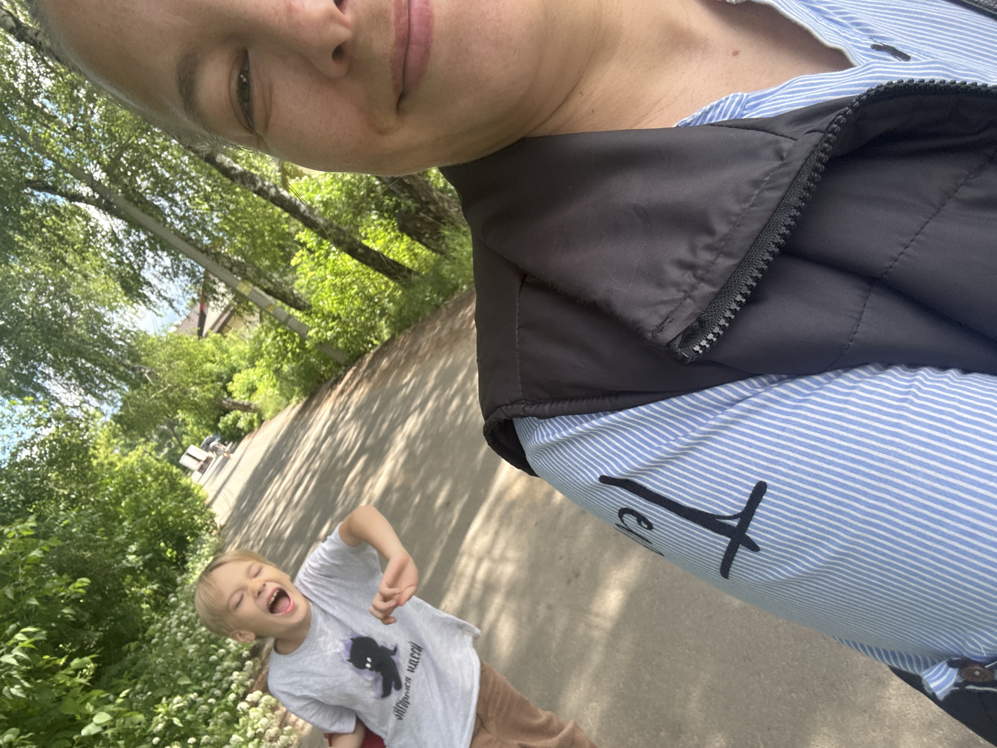 A blond boy with his mouth open in a yell or laugh is captured in a selfie-style shot taken by a woman whose face is partially visible in the foreground. The boy is wearing a grey t-shirt with a black graphic and text, and brown pants. He is on a paved path lined with trees and greenery, suggesting an outdoor, possibly park-like setting. The woman, wearing a blue and white striped shirt and a dark vest, is holding the camera and smiling at it. The path appears to be in a residential area, with glimpses of houses and more trees in the background. This scene is located in the city of Dmitrov, Russia.