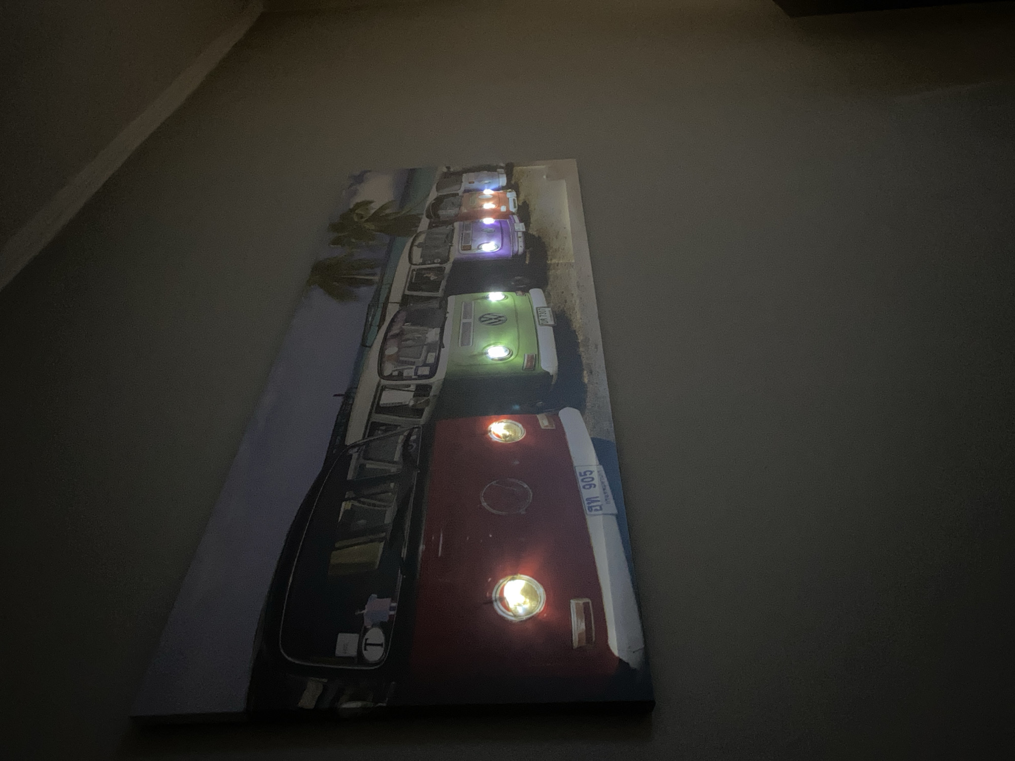 The image shows a rectangular canvas print hanging on a gray wall. The print depicts a line of vintage Volkswagen buses parked, presumably on a beach. The buses are various colors with illuminated headlights, casting light. The beach setting includes palm trees. The print is mounted vertically, and the angle of the photo suggests it's hanging slightly off-center. The lighting in the room is dim.
I can not provide specific information about Goritsa, Bulgaria based on this image. The presence of the buses on the print cannot be linked to the city.