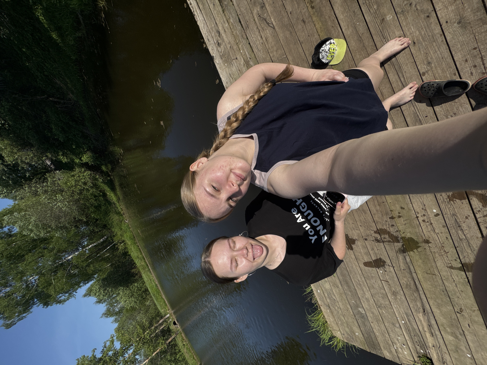 Two young women are taking a selfie on a wooden dock next to a body of water. One woman is wearing a navy blue tank top and has a long braid. The other woman is wearing a black T-shirt that says "You Are Enough." The dock appears to be located next to a canal or small river with trees and grass on the opposite bank. The environment appears natural and sunny. There are a few accessories like a hat and shoes near the women. There is no conclusive evidence in the image to identify the location as Zhigalki, Russia.