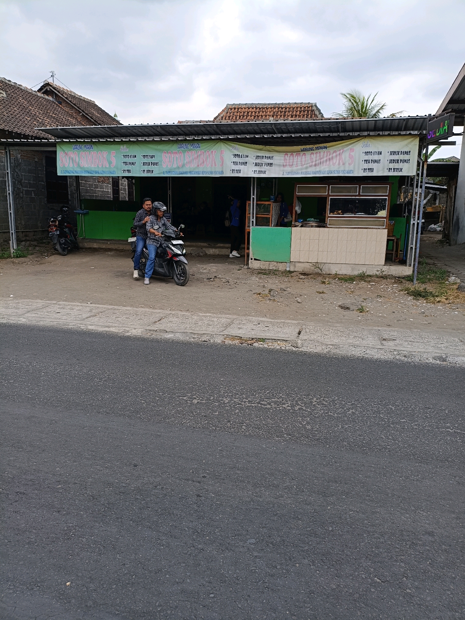 The image shows a street view of a small food stall named "SOTO SIMBOK 5" in Maguwoharjo, Indonesia. A banner above the stall lists menu items such as "SOTO AYAM," "SOTO SAPI," "TEH PANAS," "JERUK PANAS," "JERUK PANAS," and "ES JERUK." The stall itself has a counter with food displayed and a tiled facade. In front of the stall, two individuals, dressed in checkered shirts and jeans, are seated on a motorcycle. One person is driving the motorcycle, and the other is a passenger. To the left of the stall, two other motorcycles are parked. The environment appears to be an outdoor setting with a paved road in the foreground and a cloudy sky above. The buildings in the background have traditional Indonesian tiled roofs.