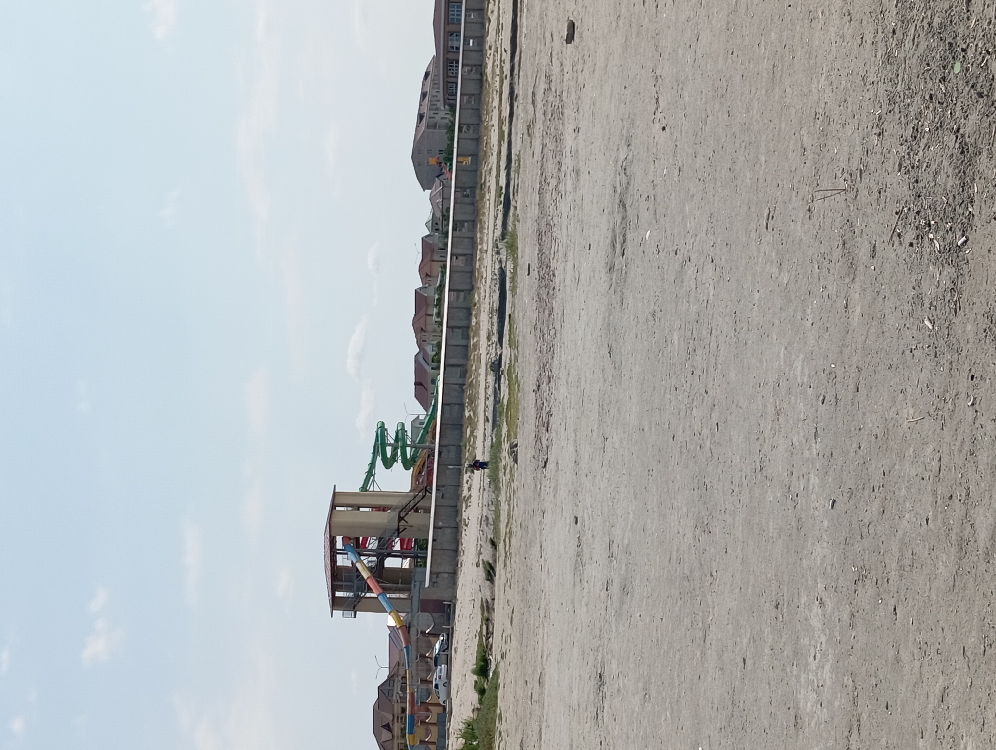 This is a wide shot of a sandy, undeveloped area with a water park in the background. In the background, a multi-story water slide structure with colorful slides is visible. To the left of the water slide, there are some houses and a few parked cars. A long, low concrete structure with openings runs horizontally across the middle ground. In the foreground, the sandy ground is mostly bare with some scattered debris and sparse vegetation. A lone figure, possibly a person, is standing in the middle ground near the concrete structure. The sky is light blue with scattered clouds. This scene is located in the city of Ömirzaq, Kazakhstan.