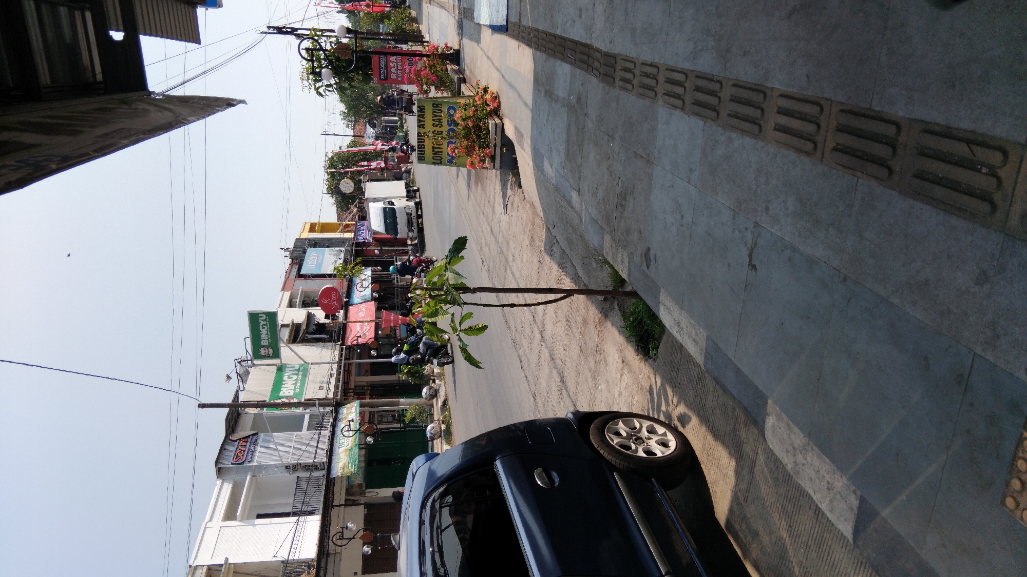 This is a street-level, eye-level medium shot of a street scene in Batang, Indonesia. The foreground is dominated by the side and rear of a dark blue sedan parked on a paved road, casting a long shadow. To the right, a concrete sidewalk is visible, with tactile paving indicating a pedestrian walkway. Small trees and potted plants with pink flowers are placed along the edge of the sidewalk.
In the mid-ground, the street is busy with a few motorcycles, some with riders wearing helmets, and a white truck. A young sapling grows on the edge of the road between the car and the sidewalk.
The background features a row of commercial buildings with storefronts, signage, and awnings. Signs for "BINGYU" (ice cream and coffee) and other businesses are visible. The sky is clear and blue, suggesting a sunny day. The overall impression is of a typical urban street with commercial activity and traffic.