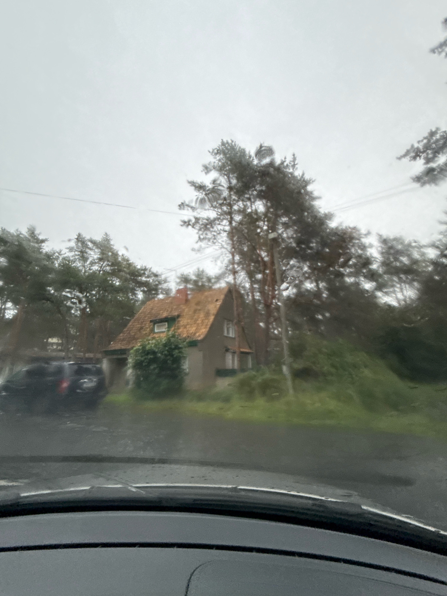 The image shows a view from inside a vehicle looking out at a house and trees. It is a cloudy and overcast day, and the windshield has raindrops on it, suggesting it is raining. The house is small with a reddish-brown tiled roof and light-colored walls. There is a bush in front of the house. To the left of the house, a dark-colored car is parked. The area is surrounded by tall trees, which appear to be pine trees. Power lines are visible in the background. The ground in front of the house and car appears wet, indicating recent or ongoing rain. There are no people visible in the image. The scene suggests a somewhat rural or suburban setting, possibly on the outskirts of a city or town. The specific location in Tallinn, Estonia, cannot be determined from the visual information provided.