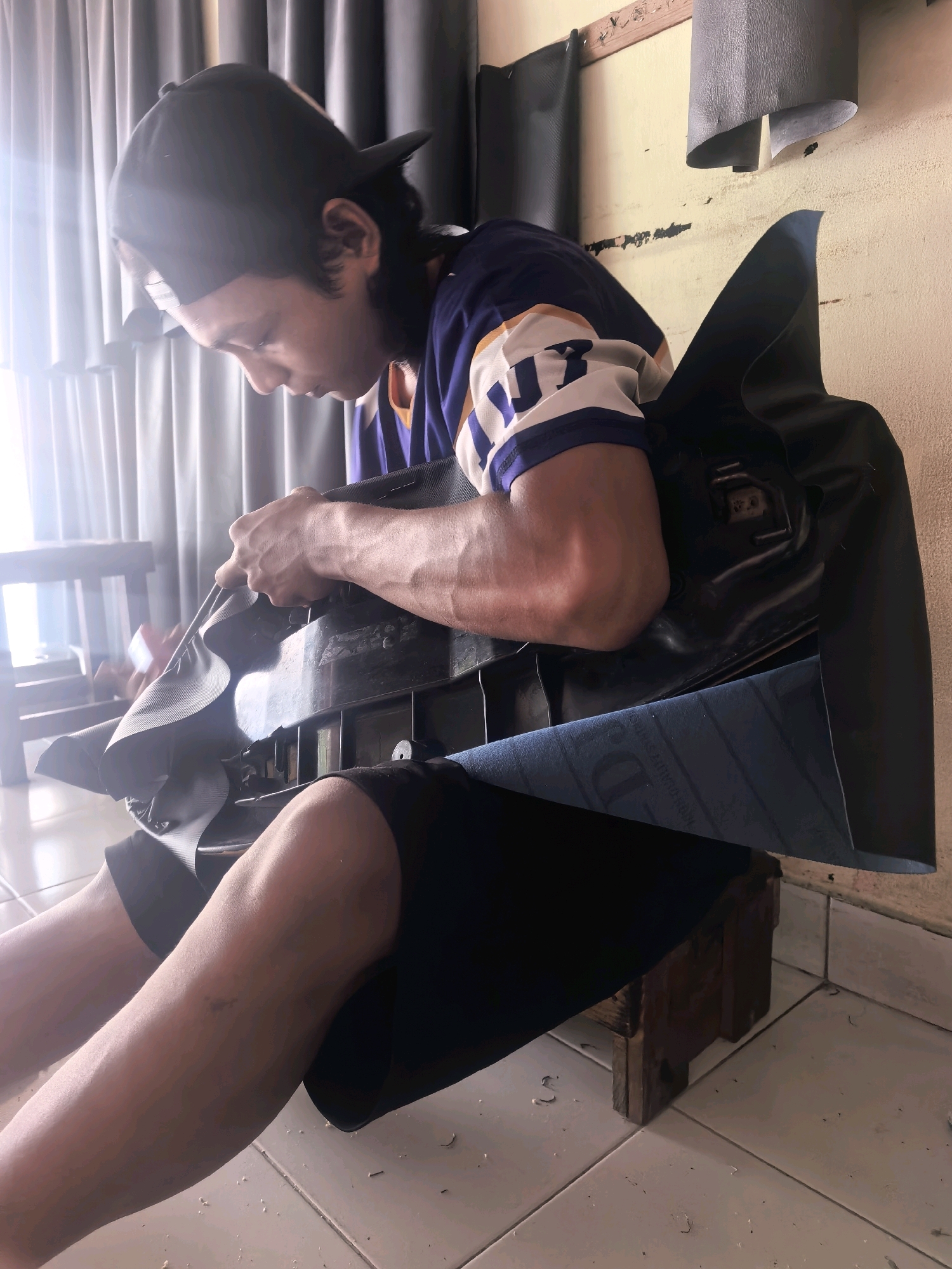 The image shows a young person sitting on the floor and working on a black object. The person is wearing a blue and white jersey with the number 17 on it and a black baseball cap. They are using a tool to shape or attach material to the black object, which appears to be a car seat cover or a similar item. A blue fabric is visible underneath the black material. The environment appears to be a workshop or a room with tiled floors, a wall with peeling paint, and some curtains in the background. The lighting suggests it is daytime, possibly with sunlight coming from the left. The overall scene conveys a sense of manual labor and crafting. The location in Kuta, Indonesia is not explicitly evident from the visual information.