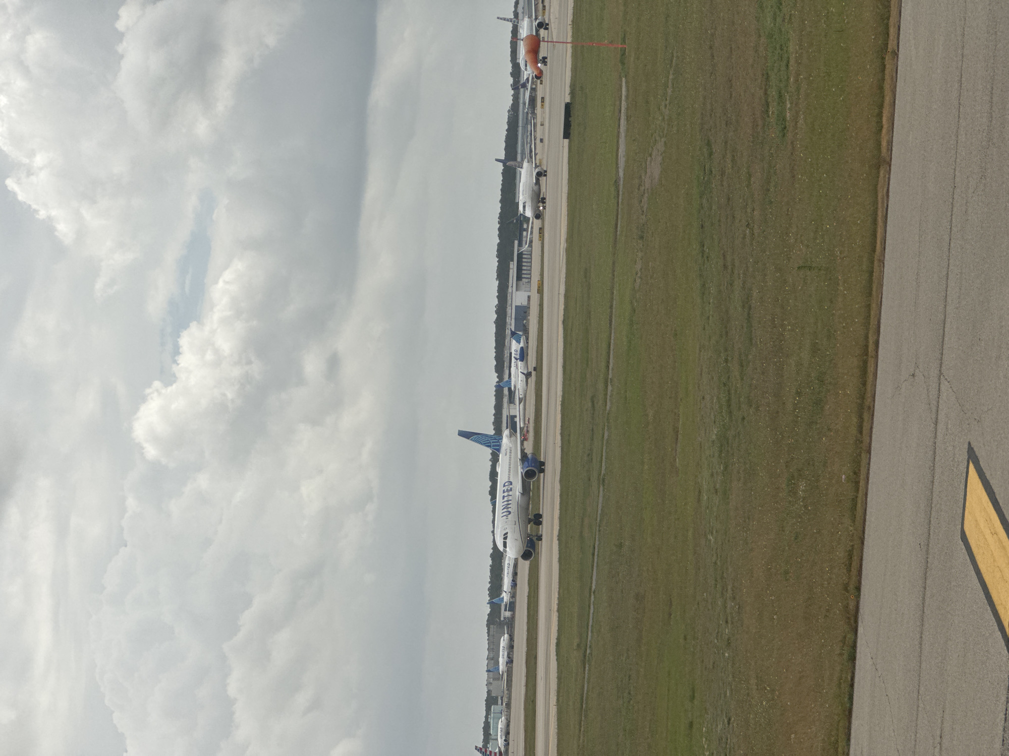 The image shows an outdoor view of an airport tarmac with airplanes aligned on the ground. One notable plane in the center displays "UNITED" on its side. The tarmac is bordered by a grassy area. Above, a cloudy sky dominates the view. The location is most likely at George Bush Intercontinental Airport (IAH) in Houston, Texas, United States of America, based on the arrangement of aircraft and the common presence of United Airlines at that airport. There are no people visible in the frame.
