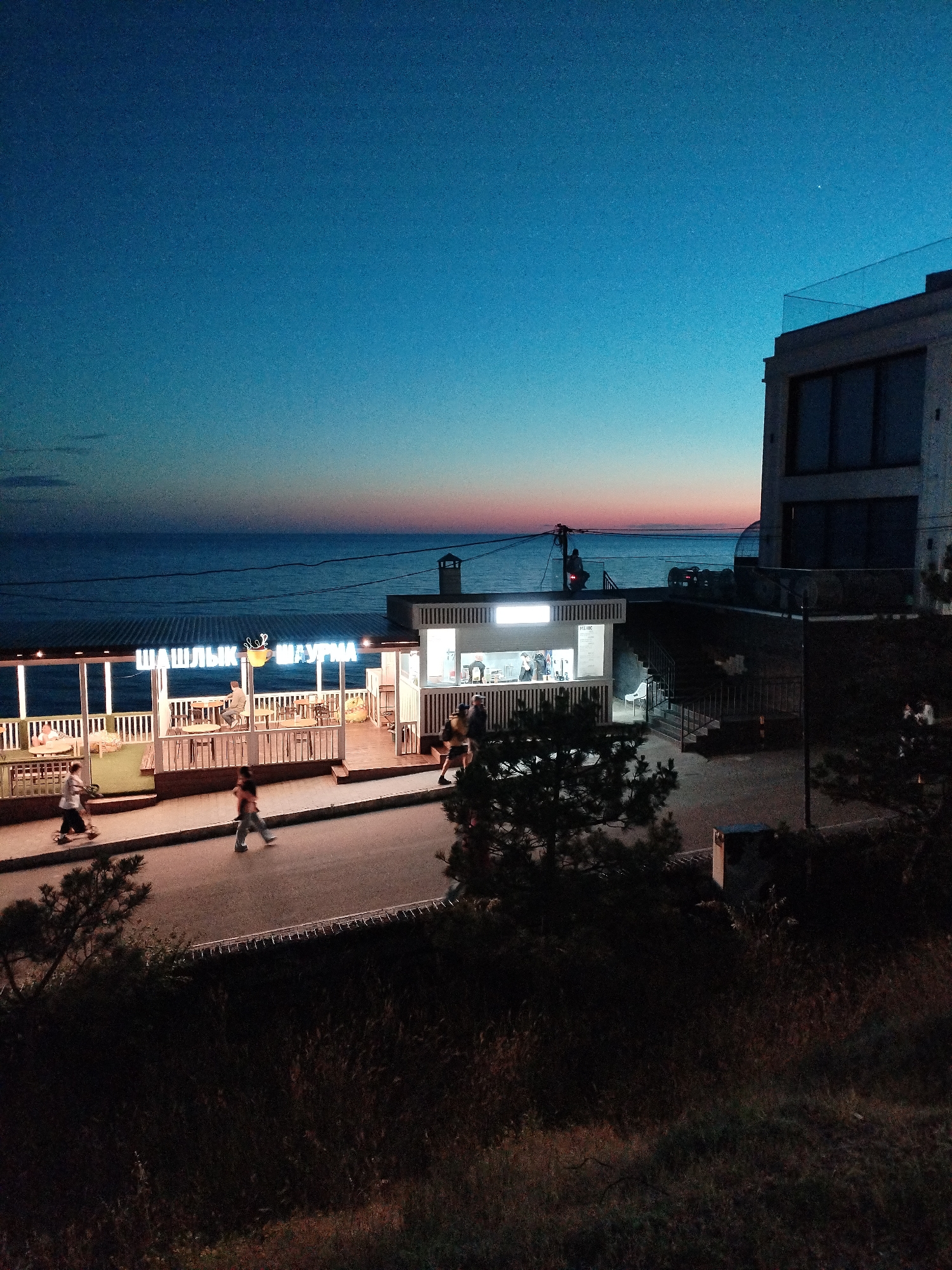 At dusk, a coastal promenade in Vinogradny, Russia, features a dining establishment with illuminated signage that reads "ШАШЛЫК ШАУРМА" (Shashlik Shawarma). The open-air seating area of the restaurant is visible, with tables and chairs set up, and several people are seated inside. In front of the restaurant, a paved walkway runs parallel to the sea, and a few individuals are walking along it. Beyond the walkway, the sea stretches to the horizon, where the sky displays a gradient of deep blue to orange and pink hues from the setting sun. To the right of the promenade, a modern multi-story building stands, its upper floors illuminated, and a glass railing adorns the rooftop. The foreground is dominated by dark, out-of-focus vegetation and a road.