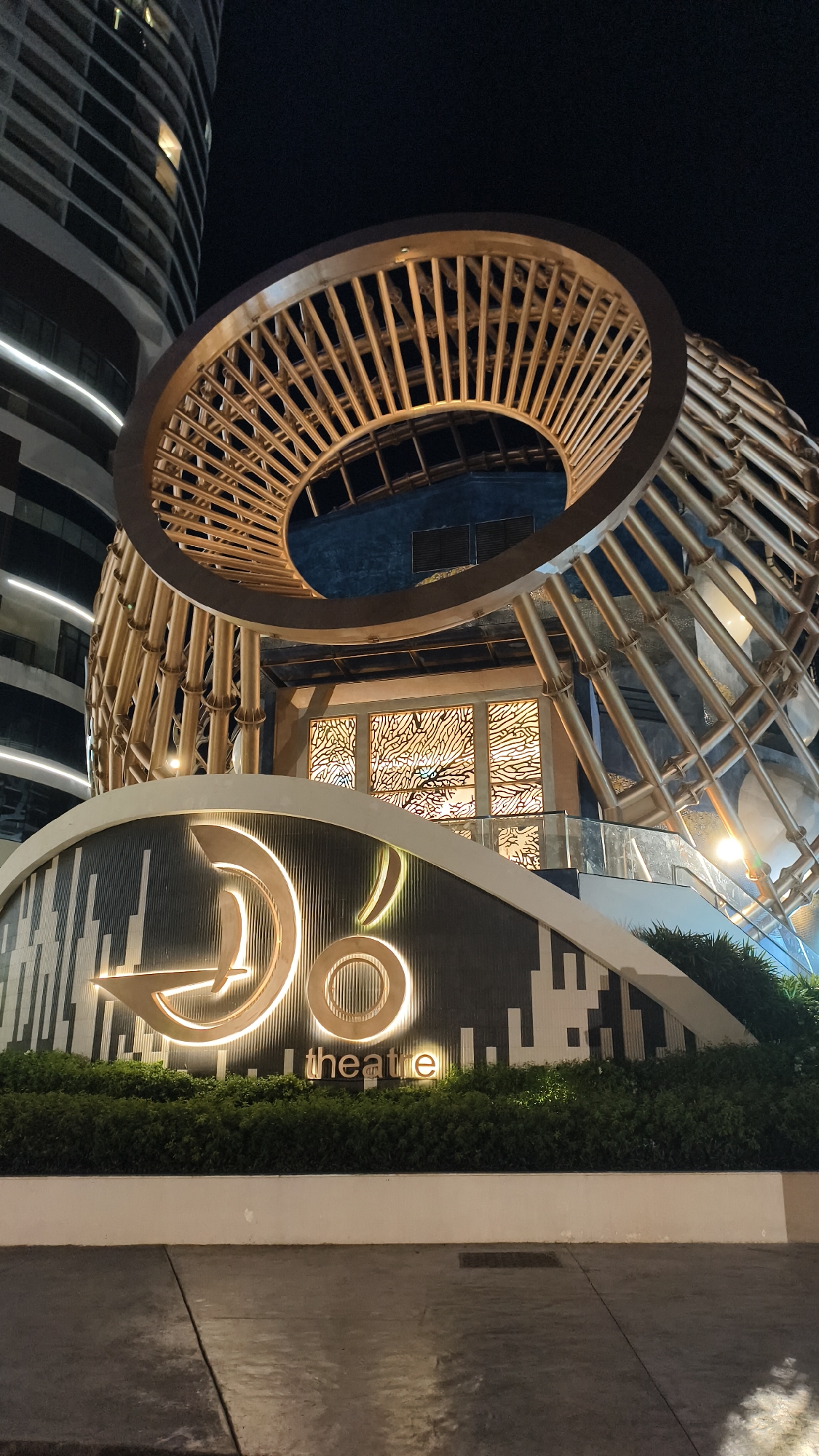 The image shows the exterior of a building at night. The building has a large, circular, metallic structure with vertical bars on its upper section, resembling a cage or a sunburst. Below this, a facade features a prominent logo with illuminated concentric circles and a stylized arrow or waveform, followed by the word "theatre" in illuminated lettering. The facade is adorned with a mosaic of black and white rectangular tiles, and the logo and "theatre" text are illuminated in a golden hue. A well-maintained green hedge runs along the base of the facade. To the left, a tall, modern skyscraper with horizontal white lines is partially visible against the dark night sky. The ground in front of the building is a paved surface, likely a sidewalk or plaza. The overall scene is well-lit, highlighting the architectural features of the building. The location is identified as Bac Nha Trang Ward, Vietnam.