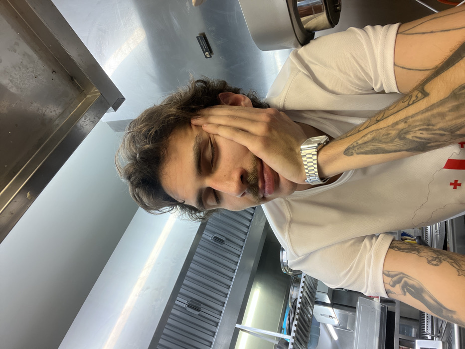 A young adult male, characterized by brown wavy hair and a moustache, is depicted in a commercial kitchen setting in Budva, Montenegro. His eyes are closed, and his right hand is positioned against his right cheek, suggesting rest or fatigue. He wears a white short-sleeved t-shirt adorned with a red cross on a white background, and a silver-toned metal wristwatch with a link bracelet on his right wrist. Visible tattoos include a large black-and-white aquatic creature on his right forearm and other designs on his left. The immediate background is dominated by stainless steel surfaces, including an industrial exhaust hood with visible grates, shelves, and overhead fluorescent lighting fixtures. Stainless steel pots are identifiable on a shelf to the left of the subject. A partially obscured dark rectangular sign, bearing the word "Montenegro," is visible on the stainless steel wall behind the subject's right shoulder. The environment is well-lit and appears to be a functional food preparation area.