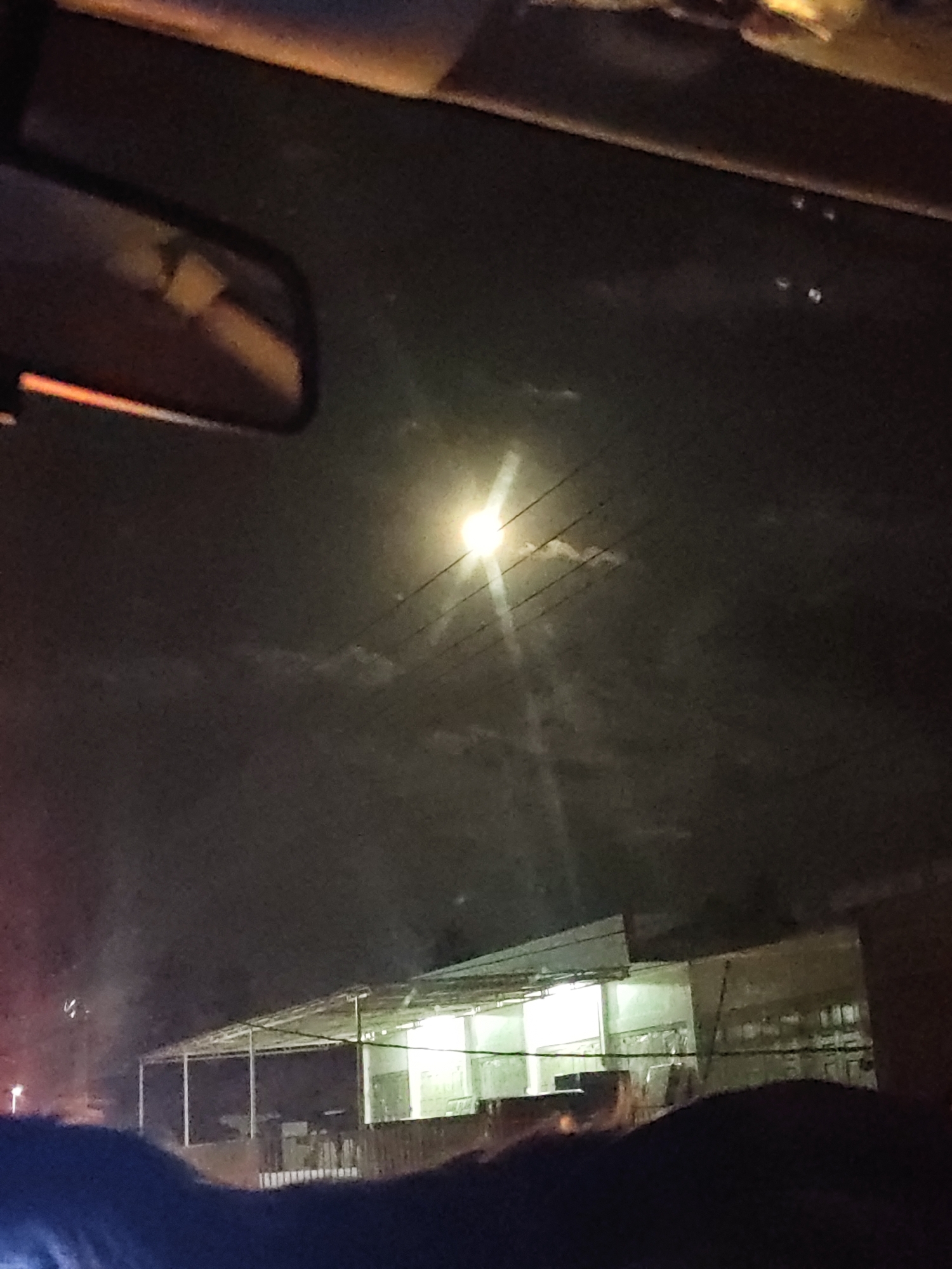 The image appears to be taken from inside a car, looking out at a street scene at night. A rearview mirror is visible in the upper left corner. A bright light source, likely a street lamp, is prominent in the center of the image, with power lines running across the sky. A building with a partially open structure is visible at the bottom of the frame. The location is presumed to be Lhoknga, Indonesia, based on the prompt. Details about people, specific objects, interactions, and the environment are limited by the image quality and viewing angle.