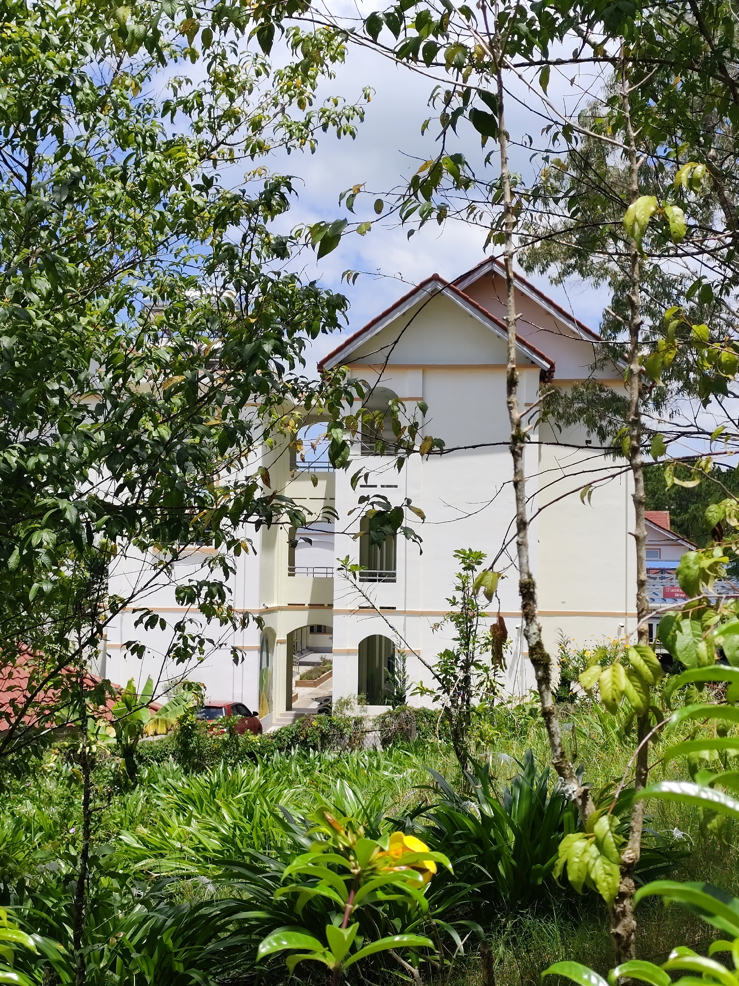 The image shows a multi-story white building with a red-tiled roof, partially obscured by lush green foliage. The building has arched windows and balconies, suggesting a residential or hotel property. The foreground is dominated by dense green vegetation, including broad-leafed plants and trees, with a bright yellow flower visible to the left. In the middle ground, a red car is parked near the entrance of the building. The background reveals a sky with scattered clouds. The scene appears to be a well-maintained property in a tropical or subtropical environment, possibly a resort or a private residence in a hilly area. Based on the architectural style and the surrounding vegetation, this scene is characteristic of many properties found in Da Lat, Vietnam, a city known for its French colonial architecture, cooler climate, and abundant pine forests and flowers.