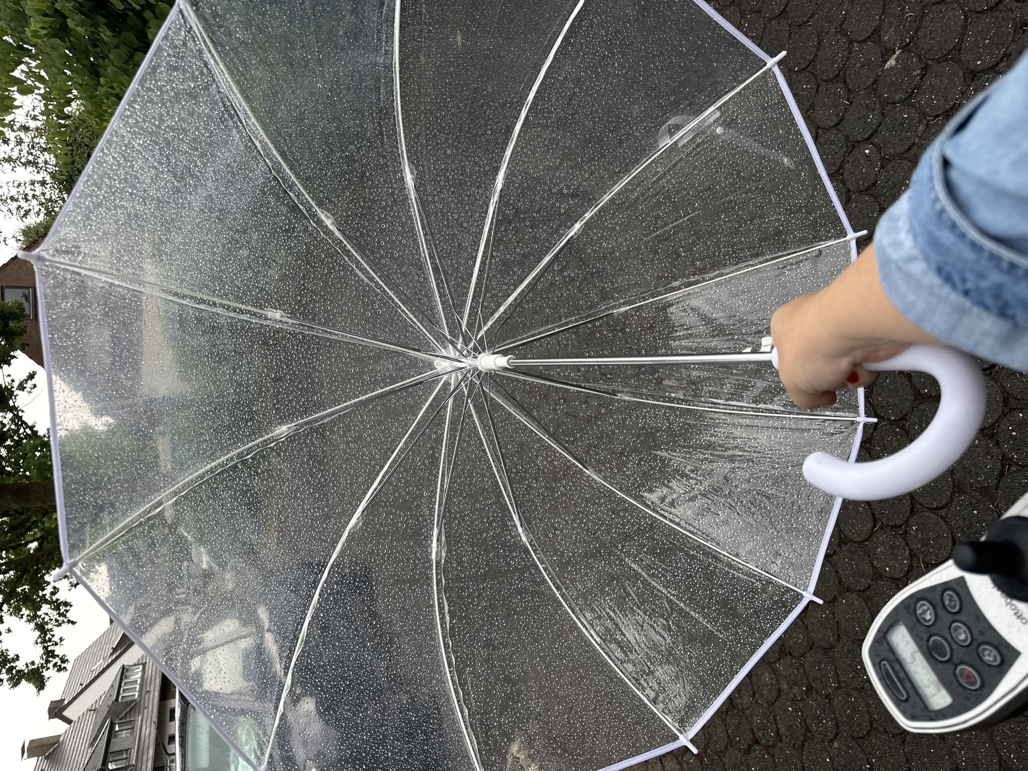 An open, clear umbrella with raindrops on it is held by a person's hand with red nail polish. The umbrella is above a black vehicle parked on a dark gray paved surface. In the lower left corner, a black and gray control unit for a mobility aid device is visible. In the background, residential buildings with tiled roofs and trees are seen under an overcast sky. The scene appears to be in a residential area of Krefeld, Germany.