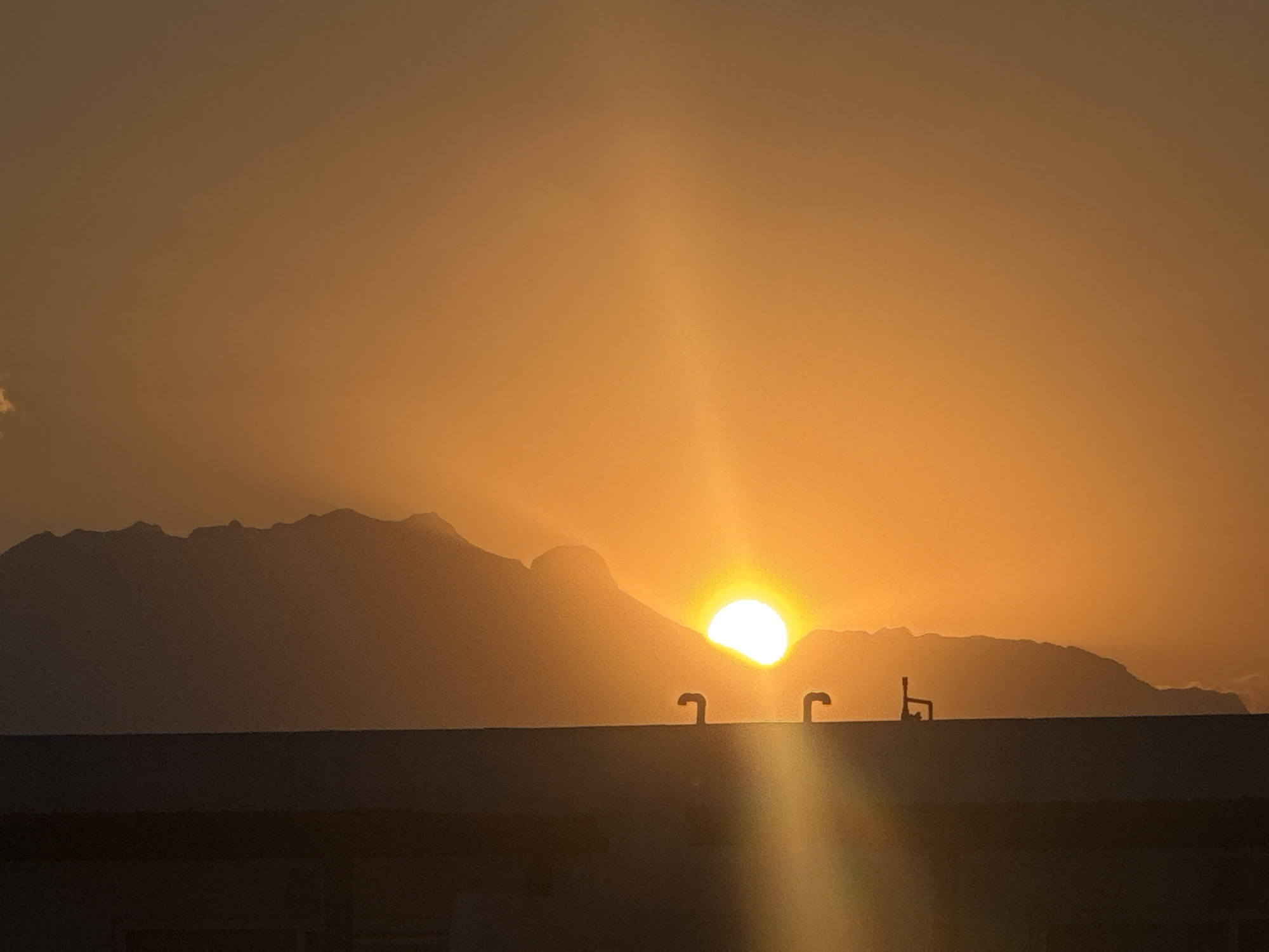 The image depicts a vibrant sunset over a mountainous landscape, with a building in the foreground, likely situated in San Francisco Ocotlán (Ocotlán), Mexico. The sky is awash in rich, warm tones of orange and yellow, suggesting either late afternoon or early evening.
The dominant feature is the setting sun, a brilliant white-yellow orb, partially obscured by the dark silhouette of a mountain range. Sunbeams radiate upwards and outwards from the sun, creating a striking glow across the sky. The mountains themselves are presented as a dark, rugged outline, showcasing their varied peaks and contours against the bright background.
In the lower portion of the frame, the dark, flat rooftop of a building extends horizontally. Two distinct, dark, pipe-like structures are silhouetted against the bright sky and sun on this rooftop. The one on the left has a pronounced curve, while the one on the right appears more angular, possibly a vent or antenna. The building itself is a dark mass, offering no details of its form or function beyond its roofline.
There is no discernible activity taking place, as it is a static landscape scene. The weather appears clear, with only a small, wispy cloud visible in the far left of the sky. No people or visible text are present in the image. The overall impression is one of peaceful natural beauty, with the dramatic light of the setting sun illuminating the scene.