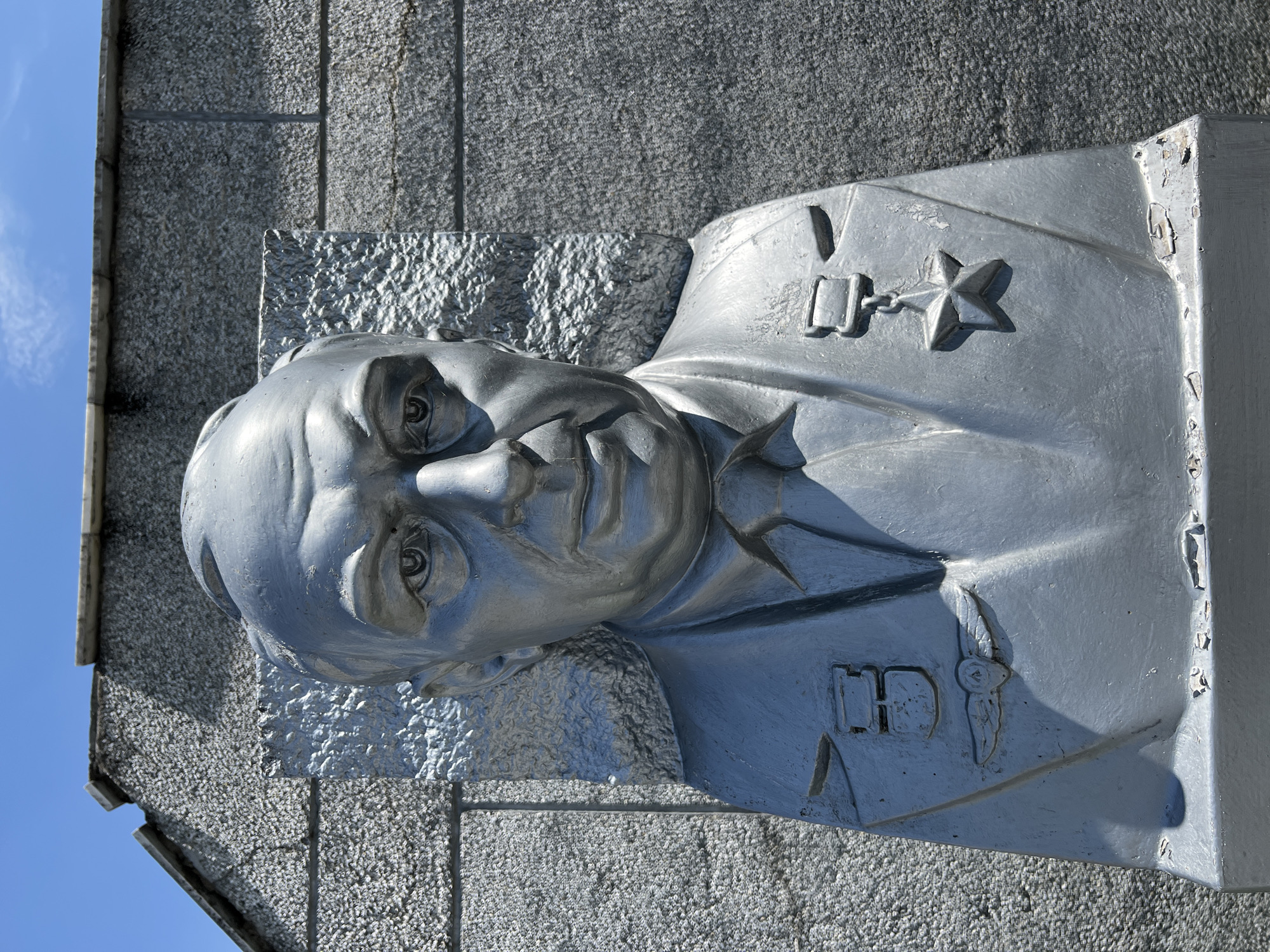 This media file shows a silver-colored bust of a man mounted on a textured stone wall. The bust is detailed, with the man depicted wearing a uniform adorned with medals. One prominent medal is a five-pointed star on a chain. The man has a serious expression, and his eyes are looking slightly downwards. The bust appears to be made of metal and shows some signs of wear and tear, with chipped paint visible on the edges.
The bust is positioned against a rough, grey stone wall, which is divided into rectangular sections by darker lines. To the left of the bust, a small portion of a rough, textured surface is visible, possibly a different type of stone or concrete. The background consists of a clear, bright blue sky.
The lighting suggests it is daytime, with the sun casting distinct shadows on the bust and the wall, highlighting the textures and contours of the sculpture. The scene appears to be outdoors, given the sky and the building materials.
The location context provided is Takhtamukai, Russia. This could be a war memorial, a monument to a historical figure, or part of a public building. There is no visible text in the image. The overall mood is solemn and perhaps commemorative, given the depiction of a decorated individual.