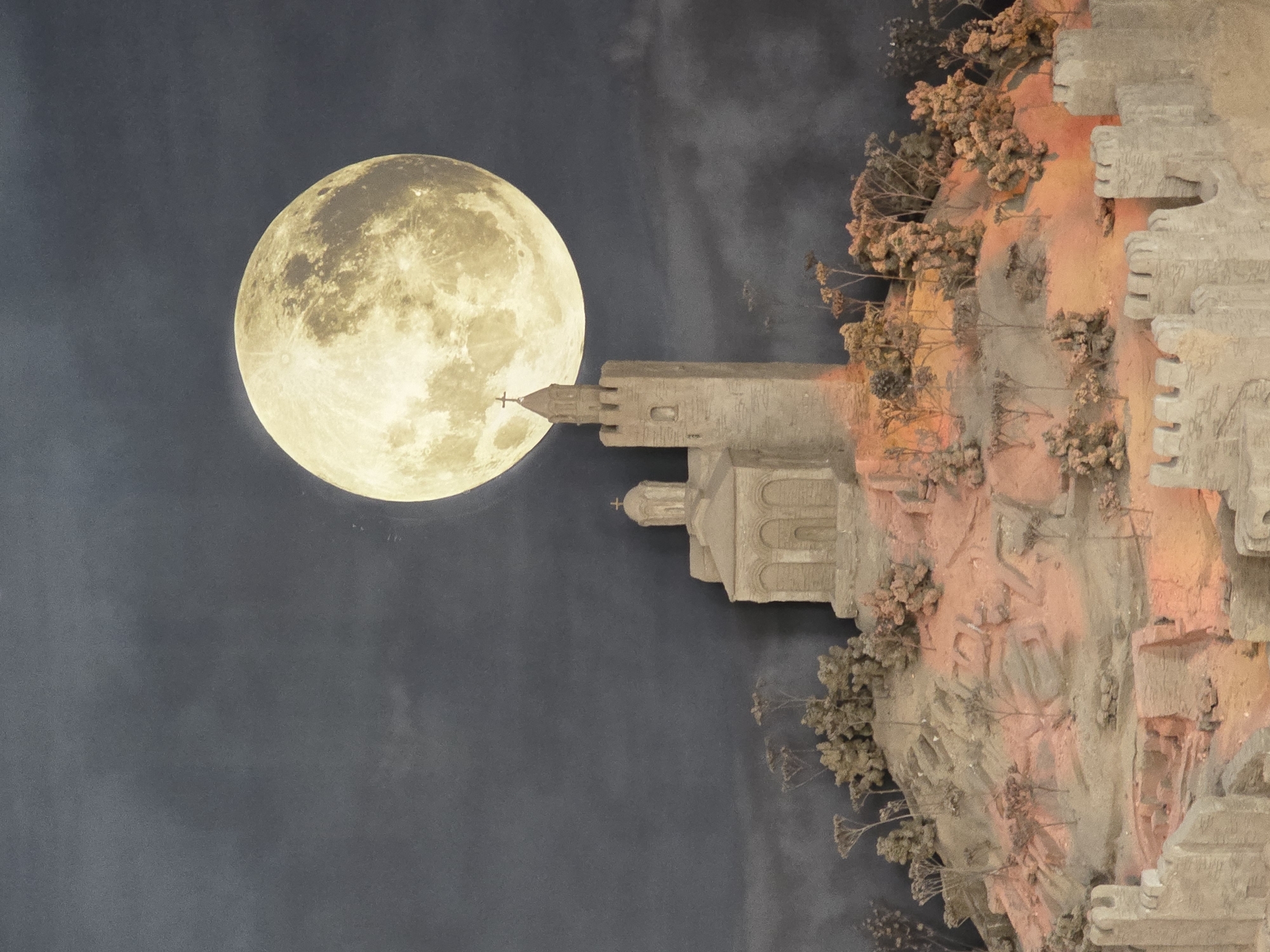 The image shows a large full moon positioned to the left of a structure resembling a medieval castle or church tower, complete with a cross on top. The tower and surrounding terrain are textured, almost like a carved stone or clay model. The "ground" adjacent to the structure is filled with what appear to be miniature trees and shrubs. The model is lit with an orange hue on its right side. The background is a dark grey, giving the impression of a night sky. The location is unknown.