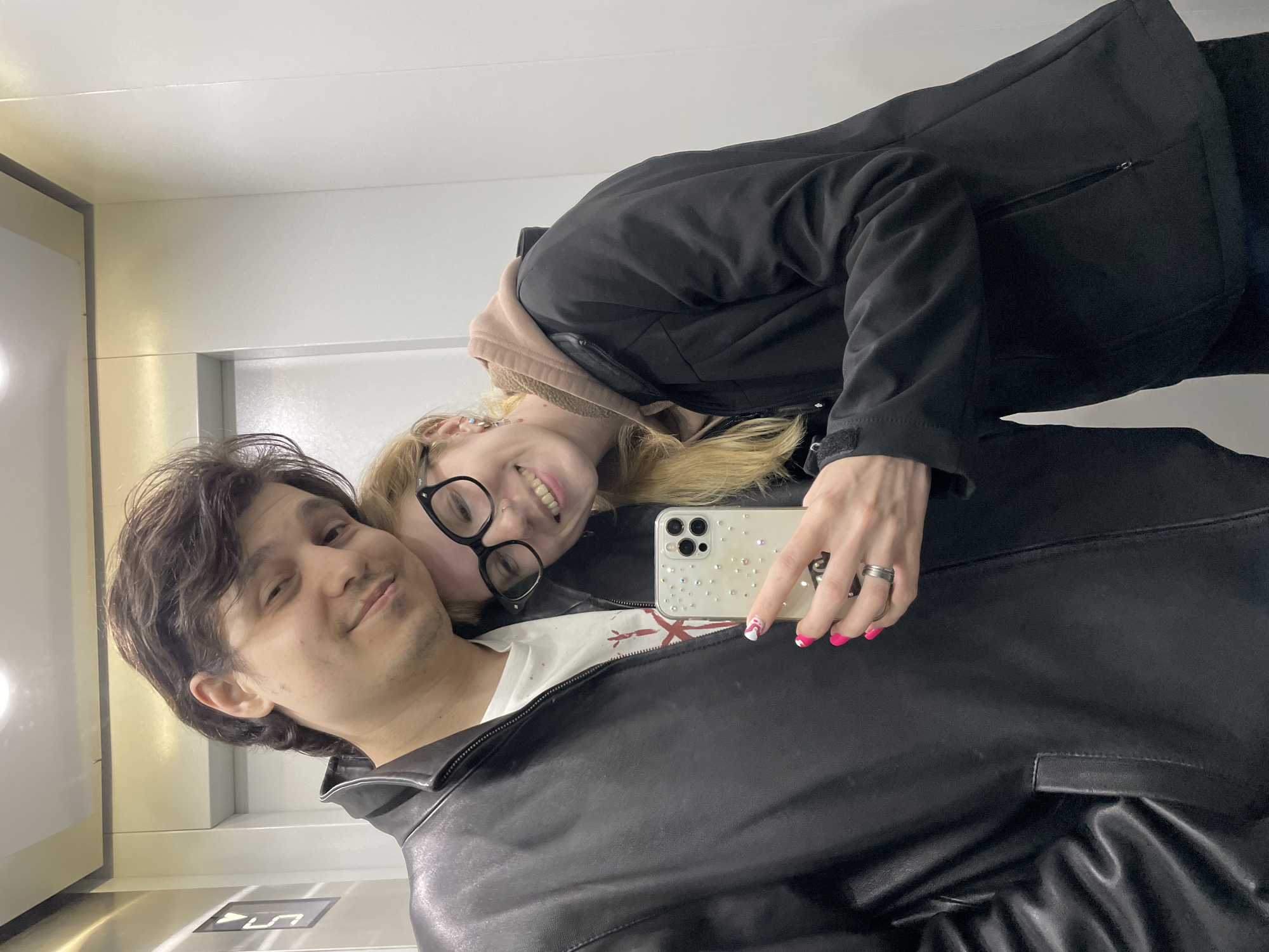A selfie captures two individuals in what appears to be an elevator or well-lit modern indoor space, likely in Izhevsk, Russia. The image is taken from a slightly upward angle, with both subjects looking towards the camera and smiling, indicating happiness and affection.
The man, positioned in the foreground and appearing to be holding the camera, has dark, wavy hair and light stubble on his face. He wears a black leather jacket over a white t-shirt with a faint red graphic design. He has a subtle smile, looking directly at the viewer.
Leaning her head on the man's shoulder is a woman with blond hair, wearing black-rimmed glasses and a nose piercing. She has a wide, cheerful smile, revealing her teeth. She is dressed in a black jacket layered over a light pink or beige hooded sweatshirt. Her nails are painted a vibrant pink with white accents. She is holding a smartphone with a clear, gem-studded case, which appears to be an iPhone, though no explicit text is clearly legible on the device itself beyond the general form factor. A silver ring is visible on her right hand.
The background consists of plain white walls and a reflective metallic strip along the left side. On the lower left, a dark floor panel is visible with illuminated symbols, including what looks like an upward arrow and possibly an alphanumeric code such as "UL" or similar, reinforcing the elevator setting. The lighting is bright and even, suggesting an indoor, artificial light source. No specific time of day or weather conditions are discernible.