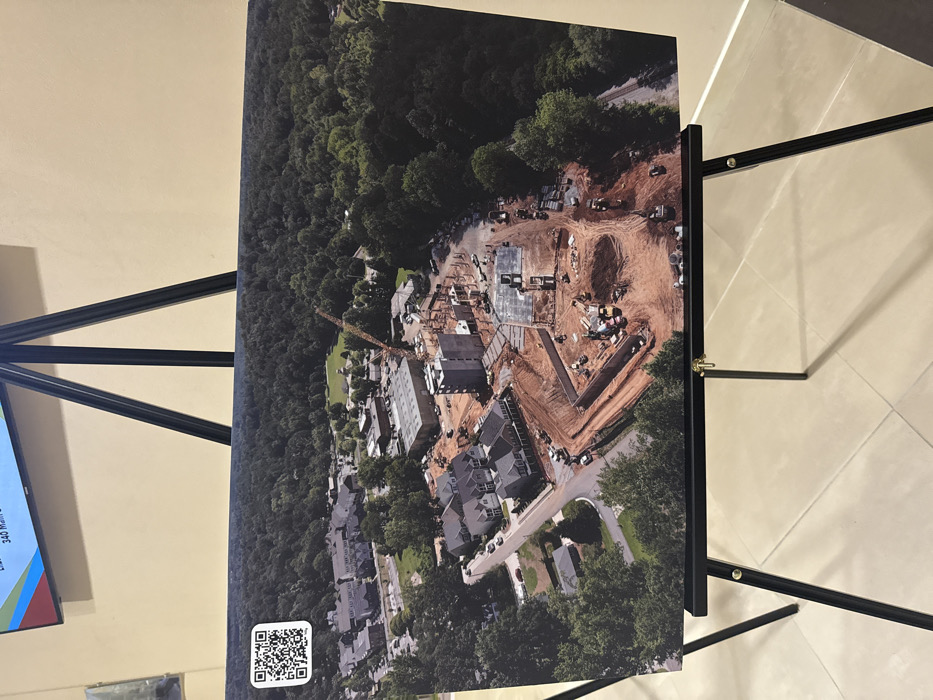 An aerial photograph depicts a construction site adjacent to a densely wooded area. A large, angular excavation forms the rough shape of a 'V', indicating potential underground construction or foundation work. Alongside, building foundations and some partially constructed structures are visible. Existing residential housing occupies the lower part of the photo, with trees interspersed between the houses and the ongoing construction. A yellow crane is also present on the site. A QR code is in the lower left.